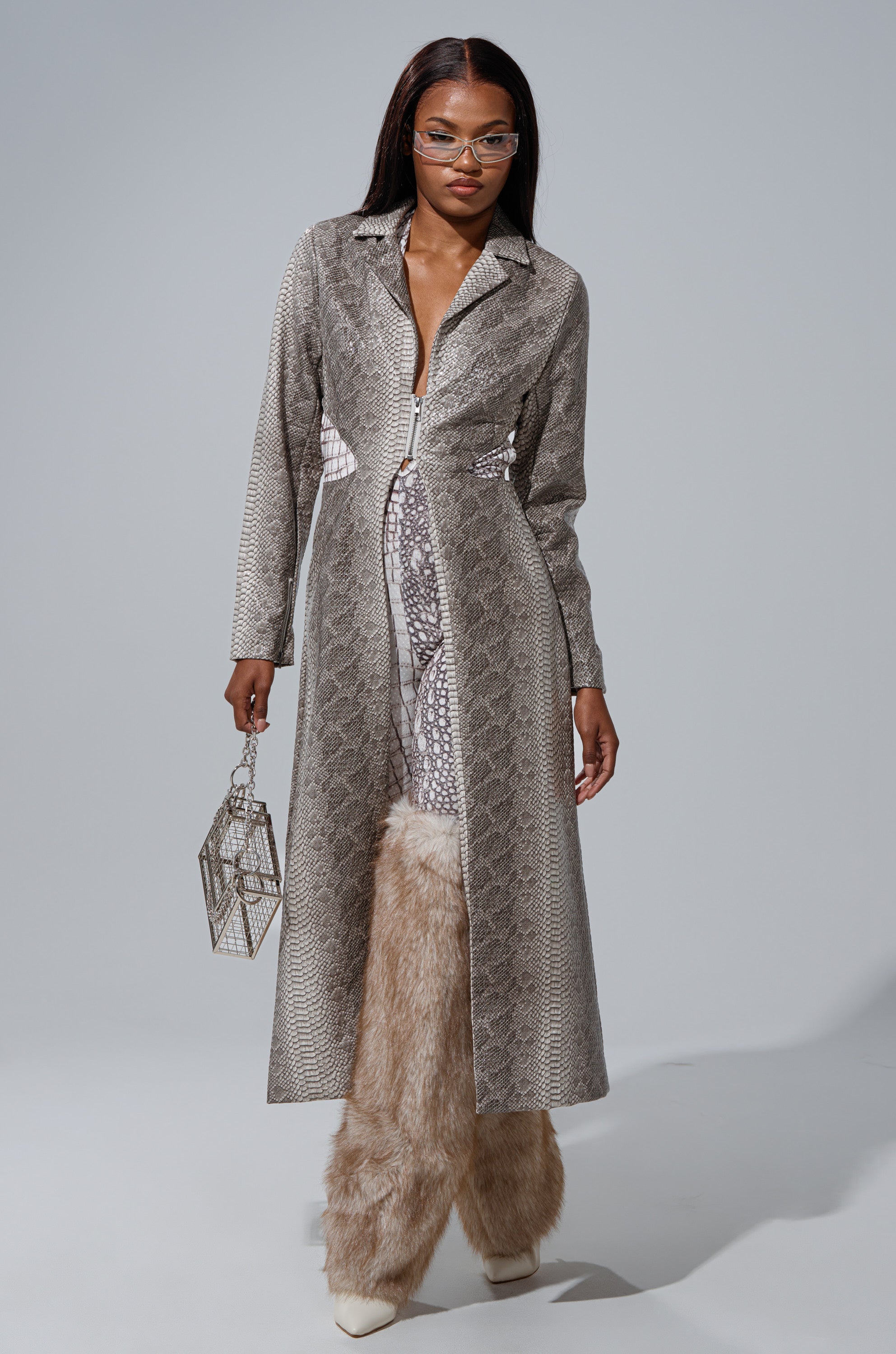 A woman stands confidently against a plain gray background wearing the LILA CUTOUT TRENCH, beige fur pants, white heels, clear-framed glasses, and holding a geometric metallic handbag. 0