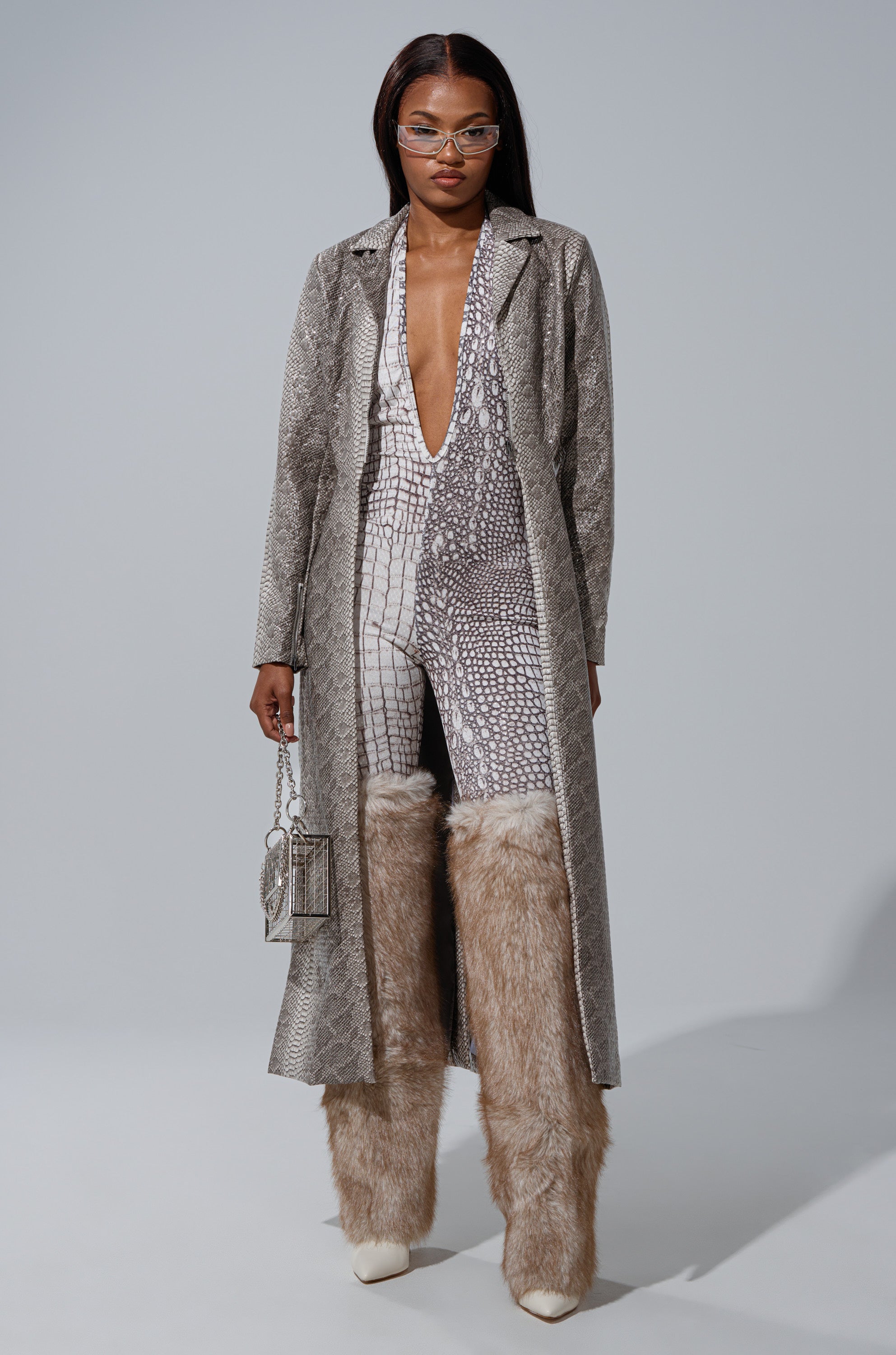 A woman in the LILA CUTOUT TRENCH, a plunging snakeskin-print jumpsuit, oversized furry beige boots, and white pointed heels stands against a gray background holding a metallic handbag. She accessorizes with clear glasses. 1