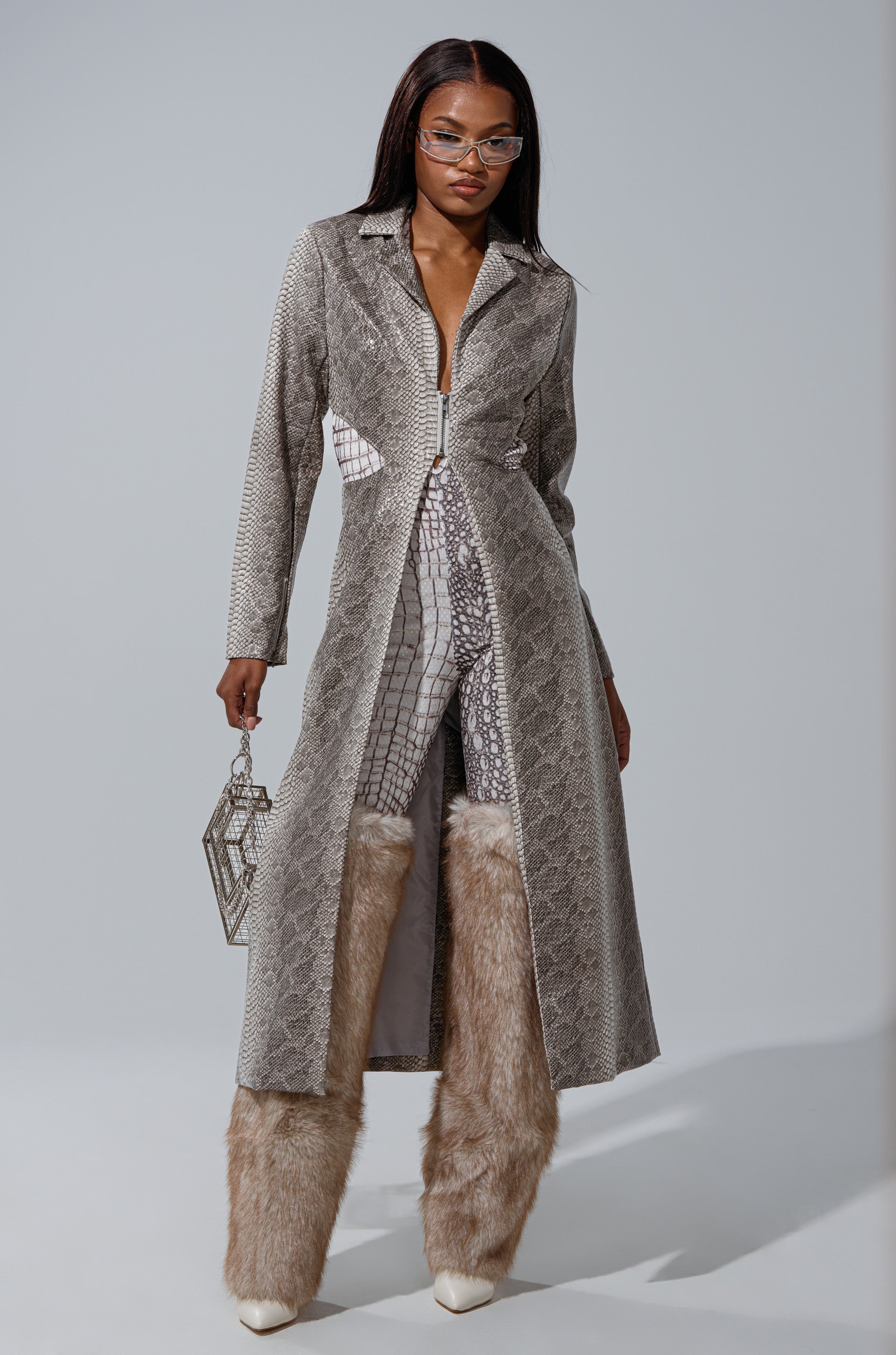 A woman in glasses models the LILA CUTOUT TRENCH, a long faux snakeskin coat layered over a matching geometric jumpsuit. She accessorizes with a chic handbag and tall, fluffy faux fur boots with pointed toes. 2