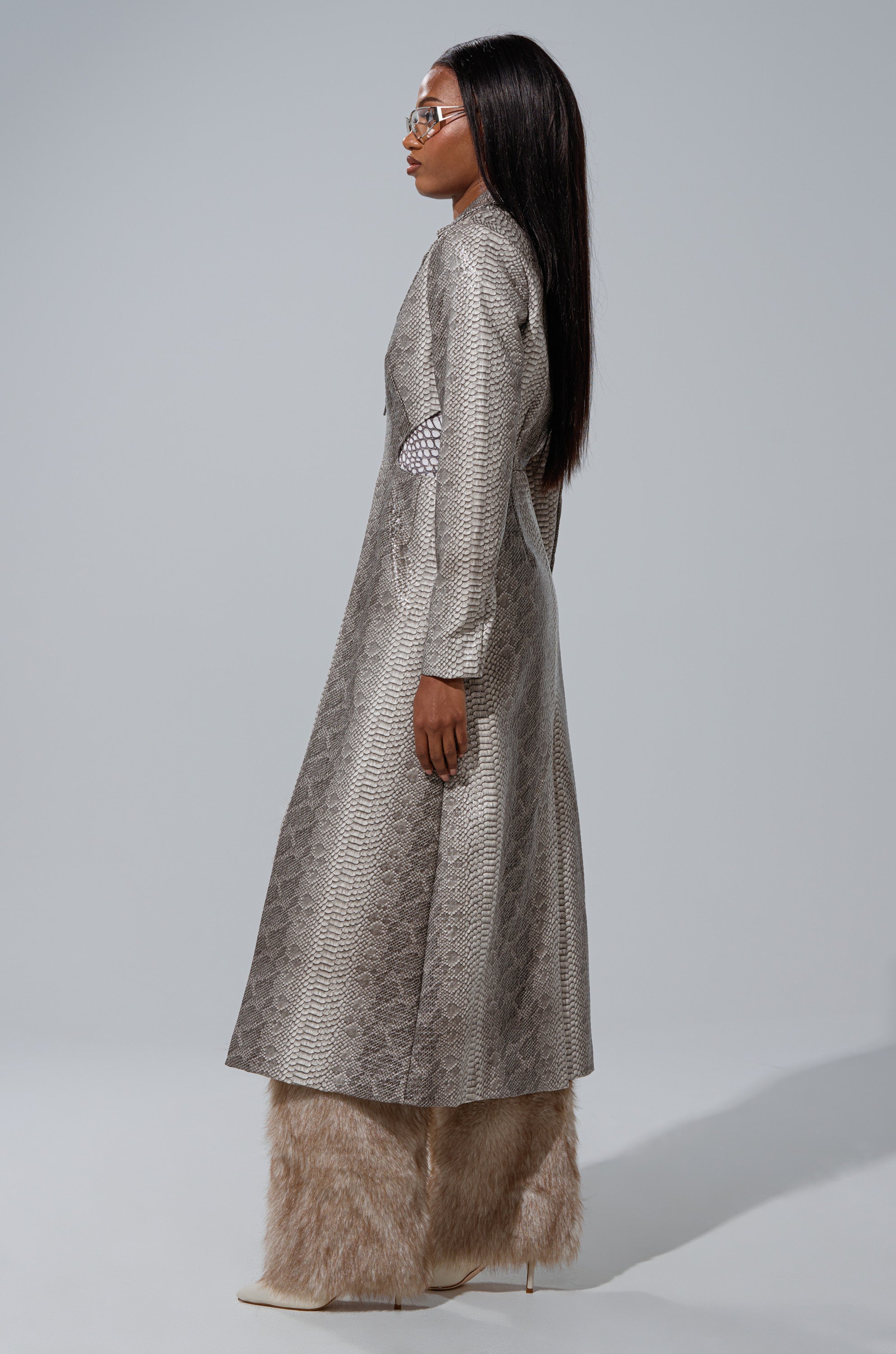A person with long straight hair and glasses poses sideways against a light gray background, wearing the LILA CUTOUT TRENCH in textured silver and beige furry pants with pointed-toe heels. 3