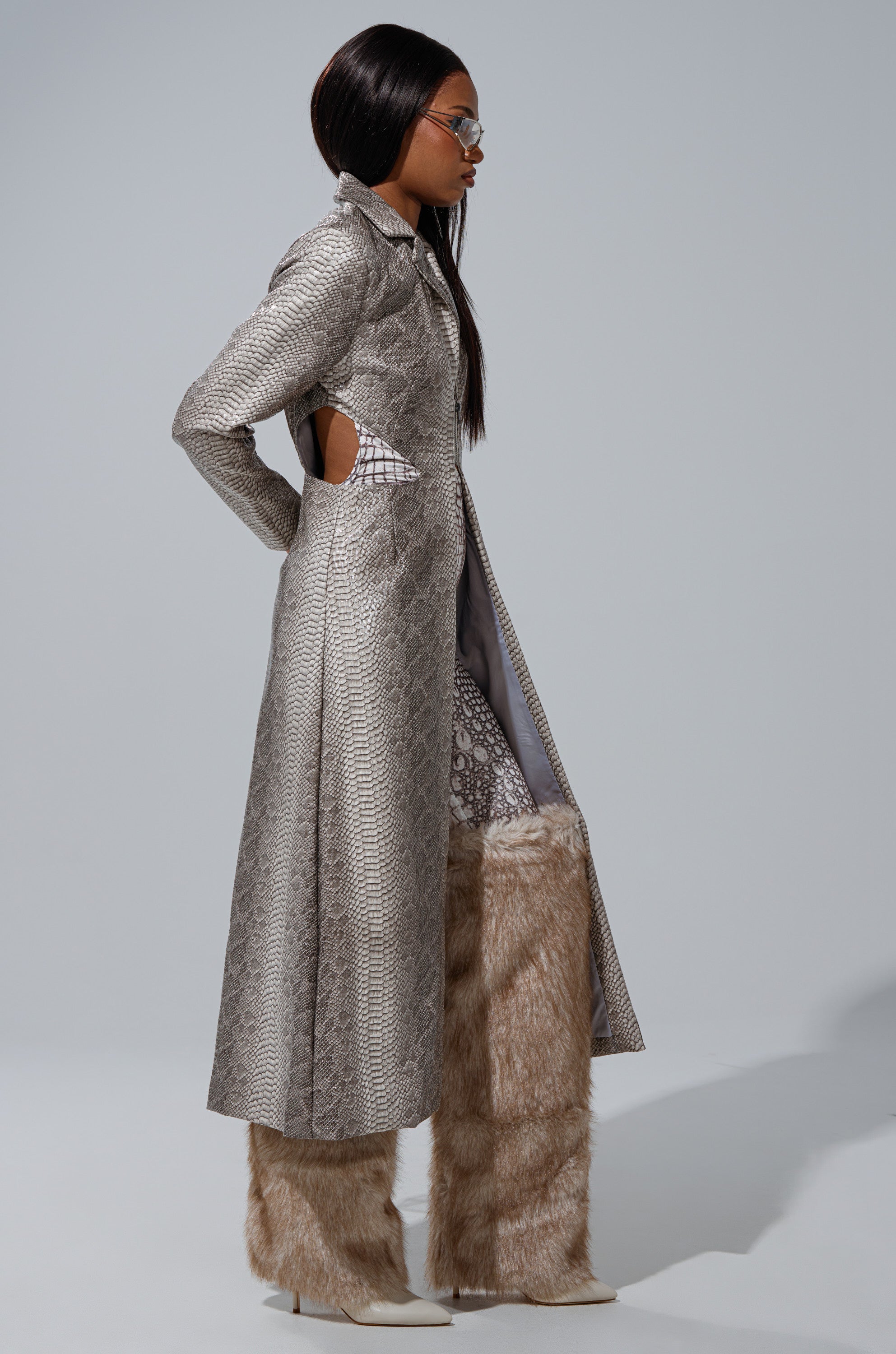 A woman models the LILA CUTOUT TRENCH, a longline faux snakeskin coat with side cutouts, layered over a patterned look and paired with tall light brown faux fur pants, white pointed shoes, and sunglasses against a plain backdrop. 4