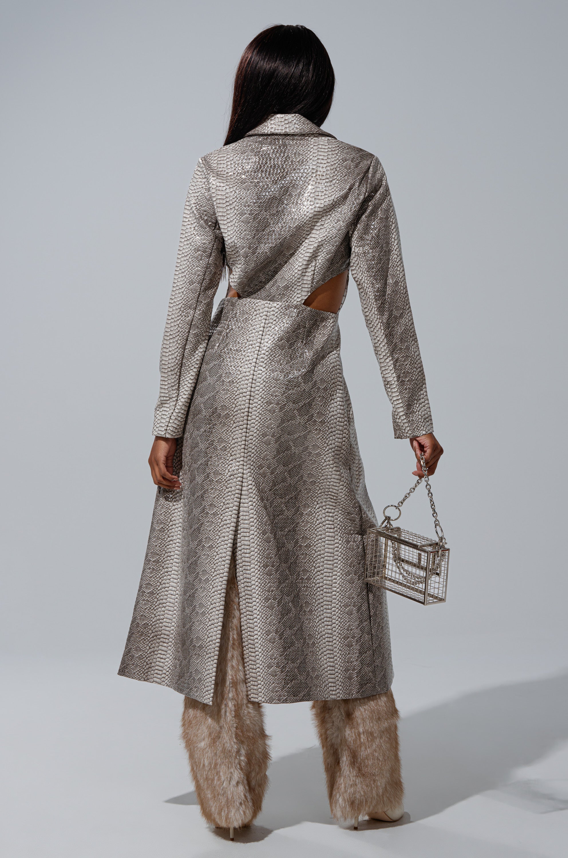 Wearing the LILA CUTOUT TRENCH in silver faux snakeskin, a person with long hair pairs it with furry beige pants and a transparent box-shaped handbag, standing with their back to the camera against a plain background. 5