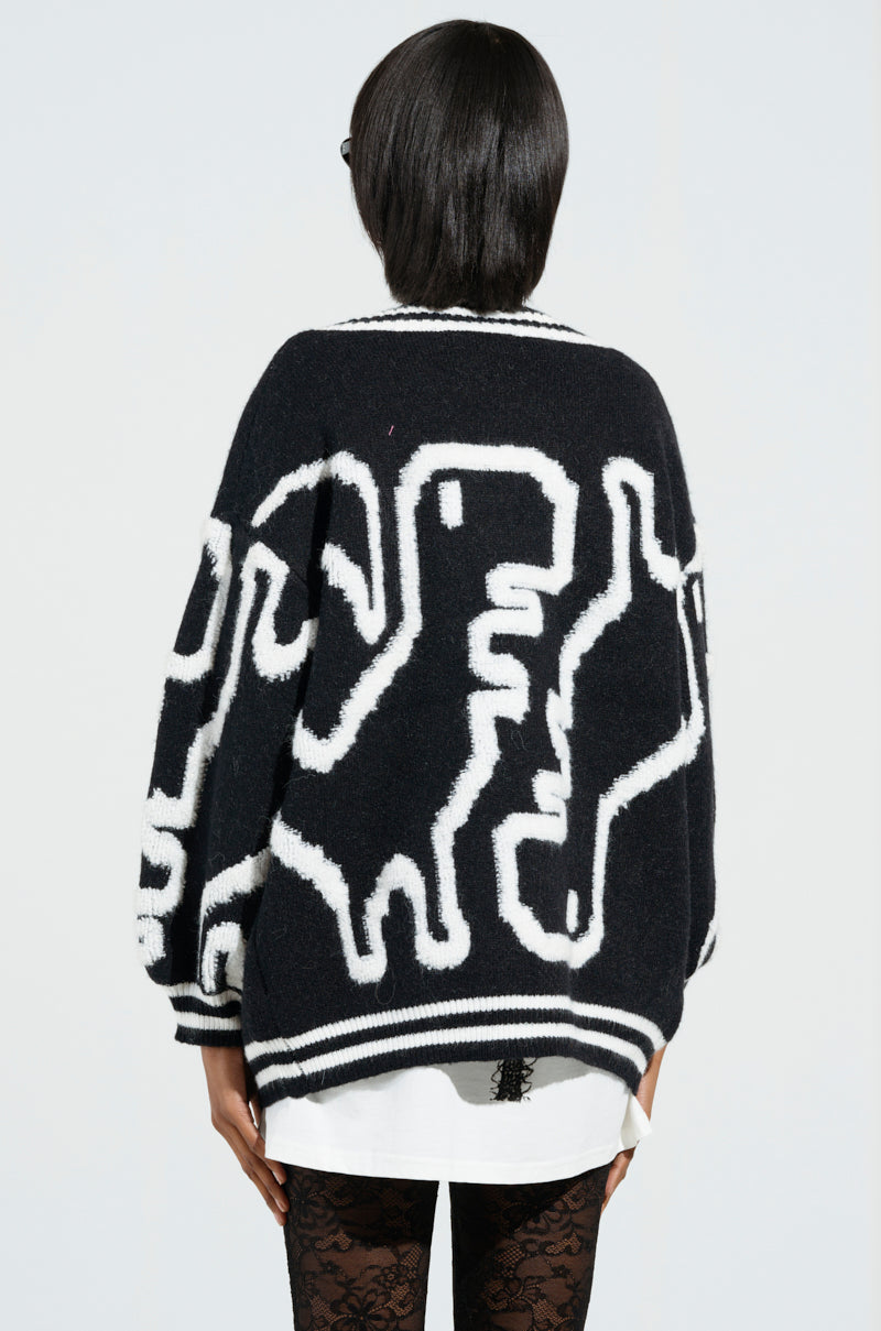 A person is seen from behind, draped in the Abstract Lover Oversized Cardigan in Black and White. This piece features abstract animal shapes over a crisp shirt, with striped cuffs and hem accentuating its stretchy knit fabrication against a plain, light-colored backdrop.