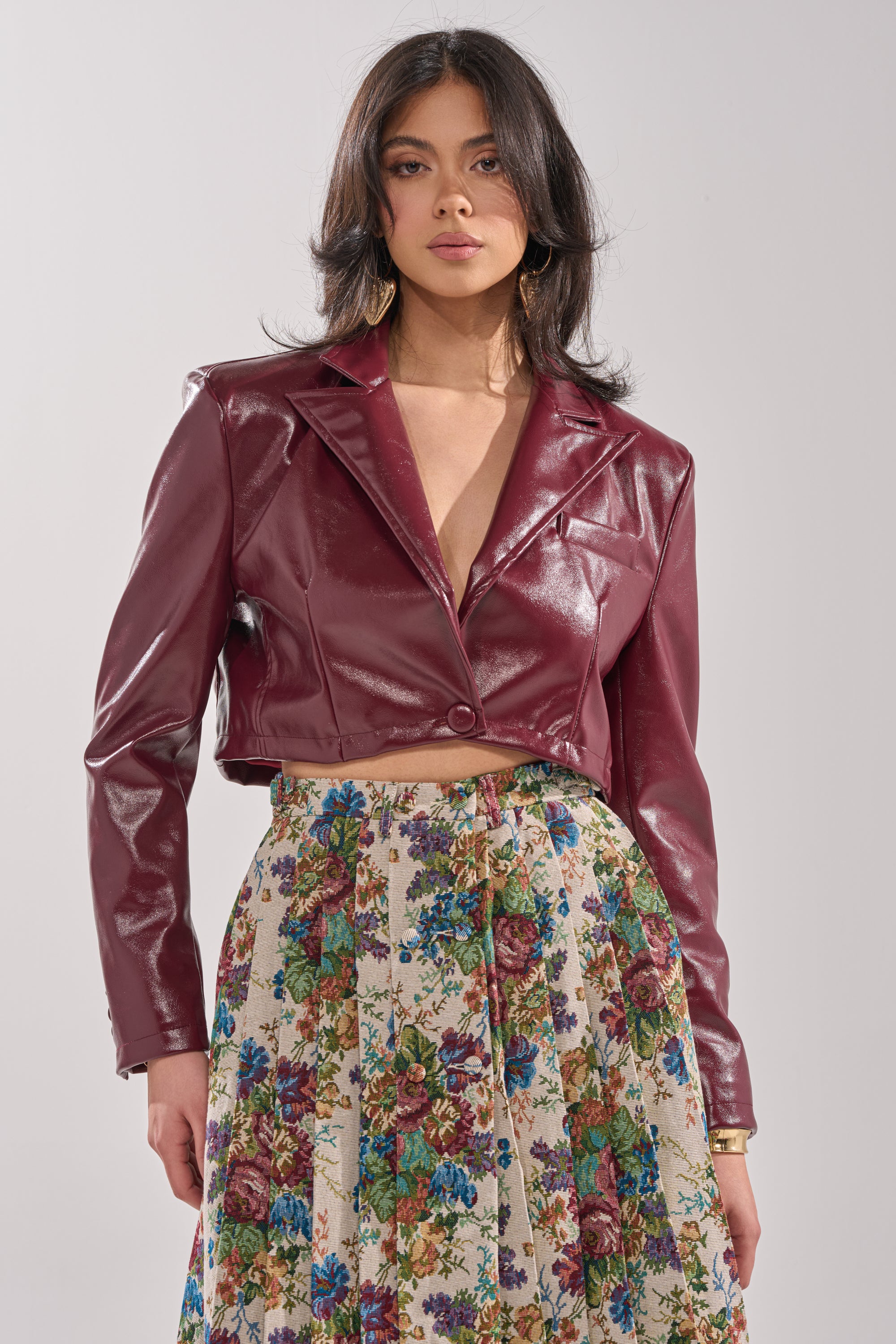 A woman wears the PENNY CROPPED FAUX LEATHER BLAZER in maroon with a tailored fit, paired with a high-waisted, pleated floral skirt, standing against a plain light background. 1