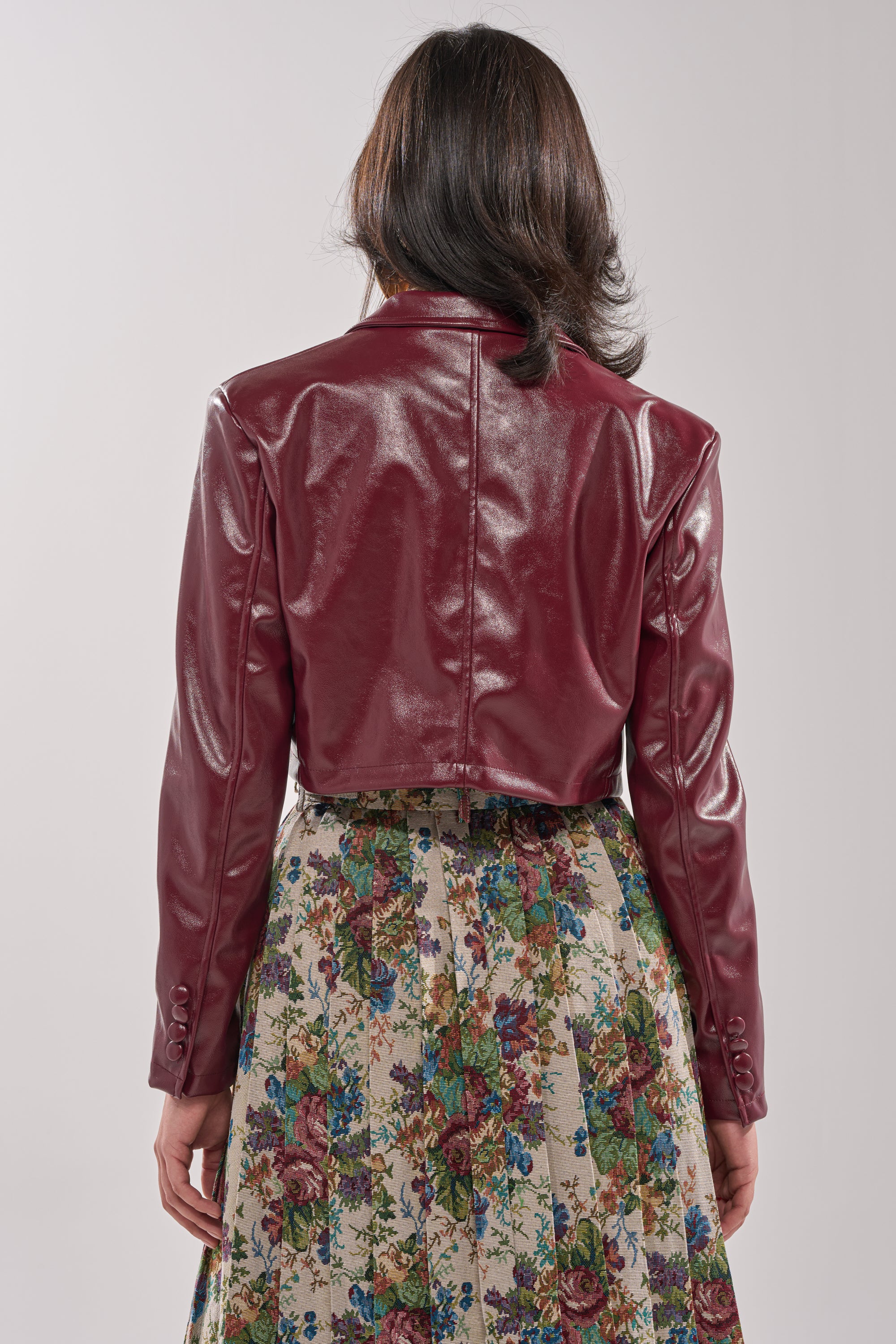 A person with shoulder-length dark hair is seen from behind, wearing the shiny maroon PENNY CROPPED FAUX LEATHER BLAZER and a high-waisted, ankle-length skirt with a colorful floral pattern. 3