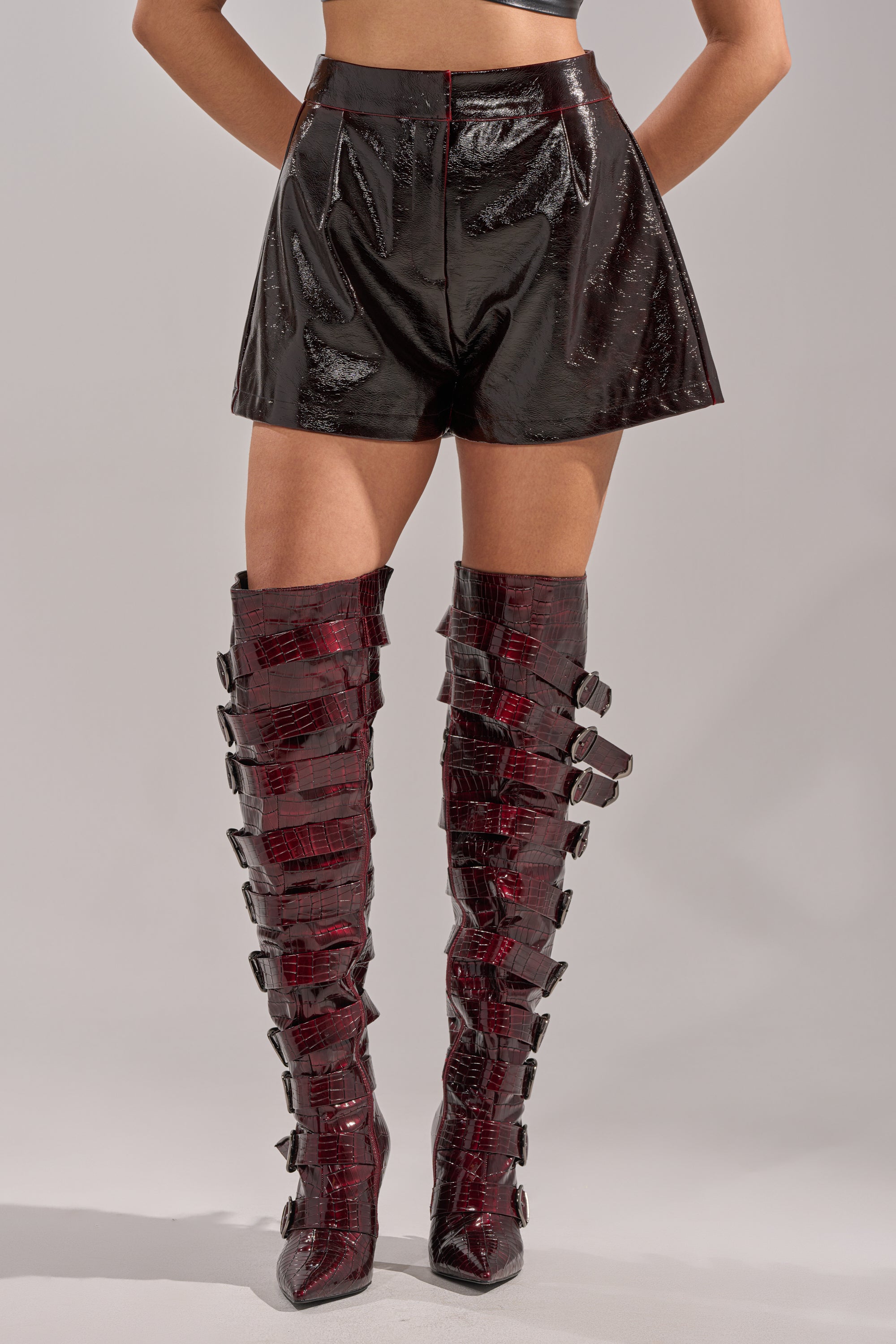 A person wearing OH HOT DAMN FAUX LEATHER SHORT black, high-waisted faux leather shorts and tall burgundy boots with multiple buckles stands hands on hips against a plain background. 2