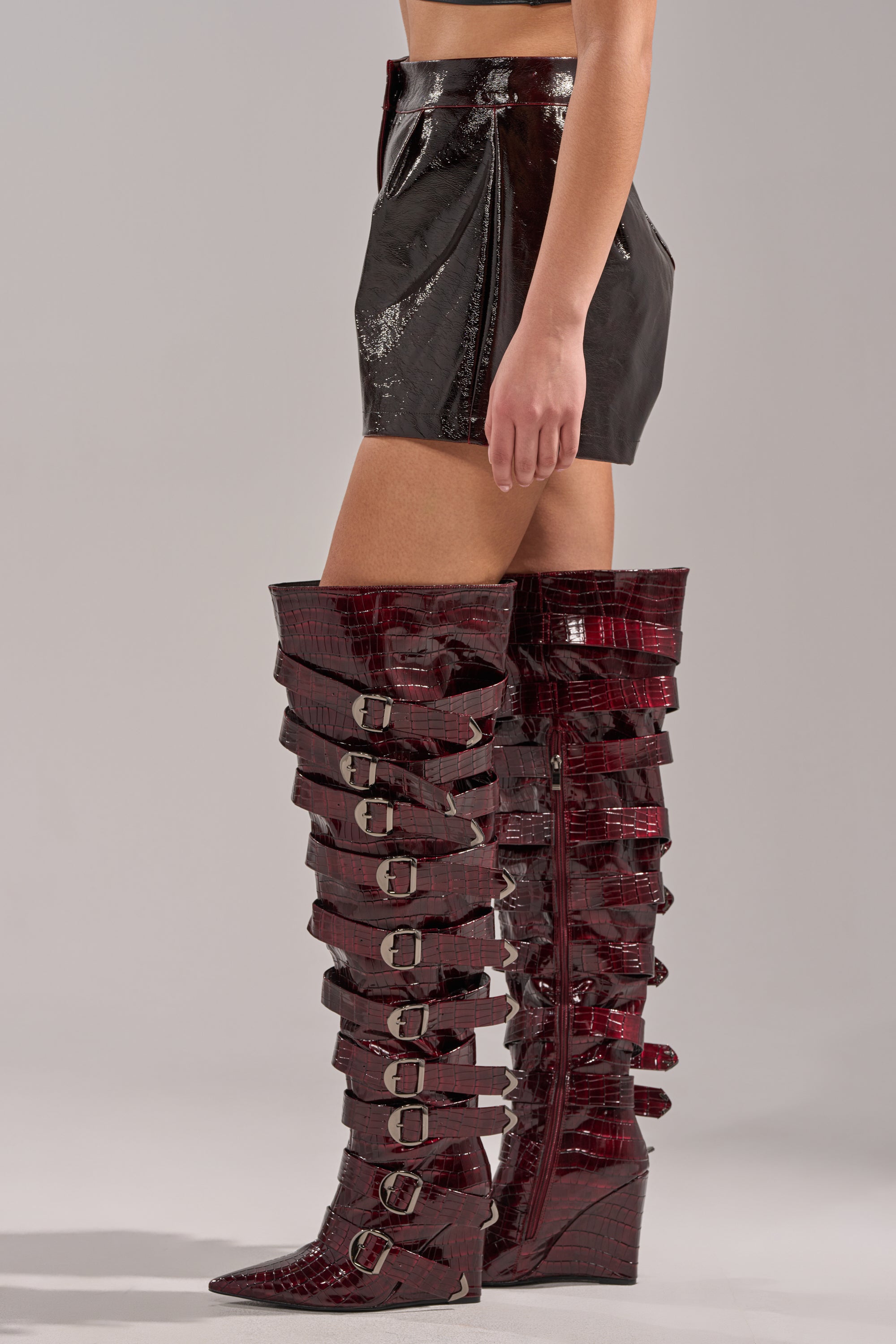 Wearing the OH HOT DAMN FAUX LEATHER SHORT, a vegan black mini skirt, paired with thigh-high, dark red crocodile-pattern boots with buckles and wedge heels, the person stands against a plain gray background. 3
