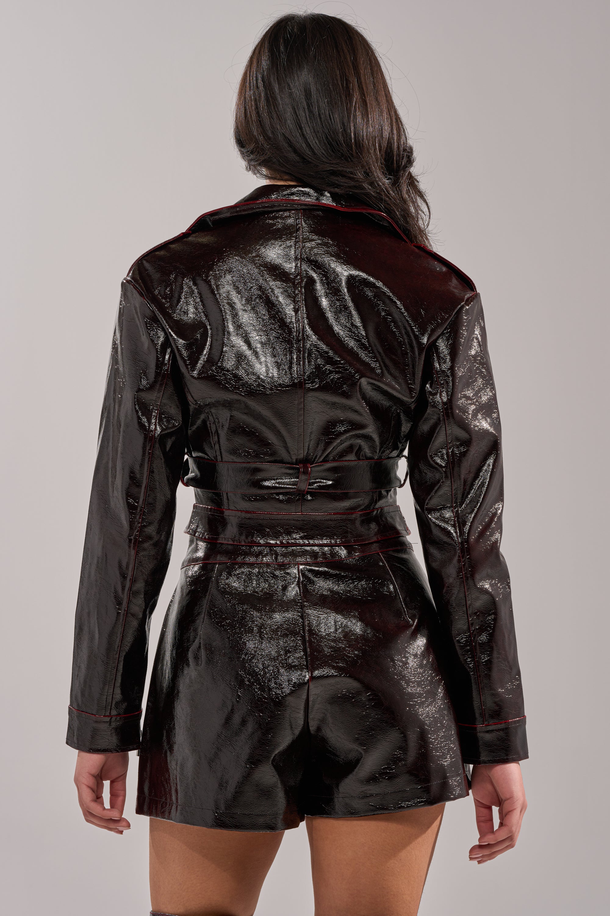 A person with long dark hair faces away, wearing THE WAY YOU MOVE COLOR CHANGING MOTO jacket and matching shorts in shiny black vegan leather. The background is plain, light gray. 4
