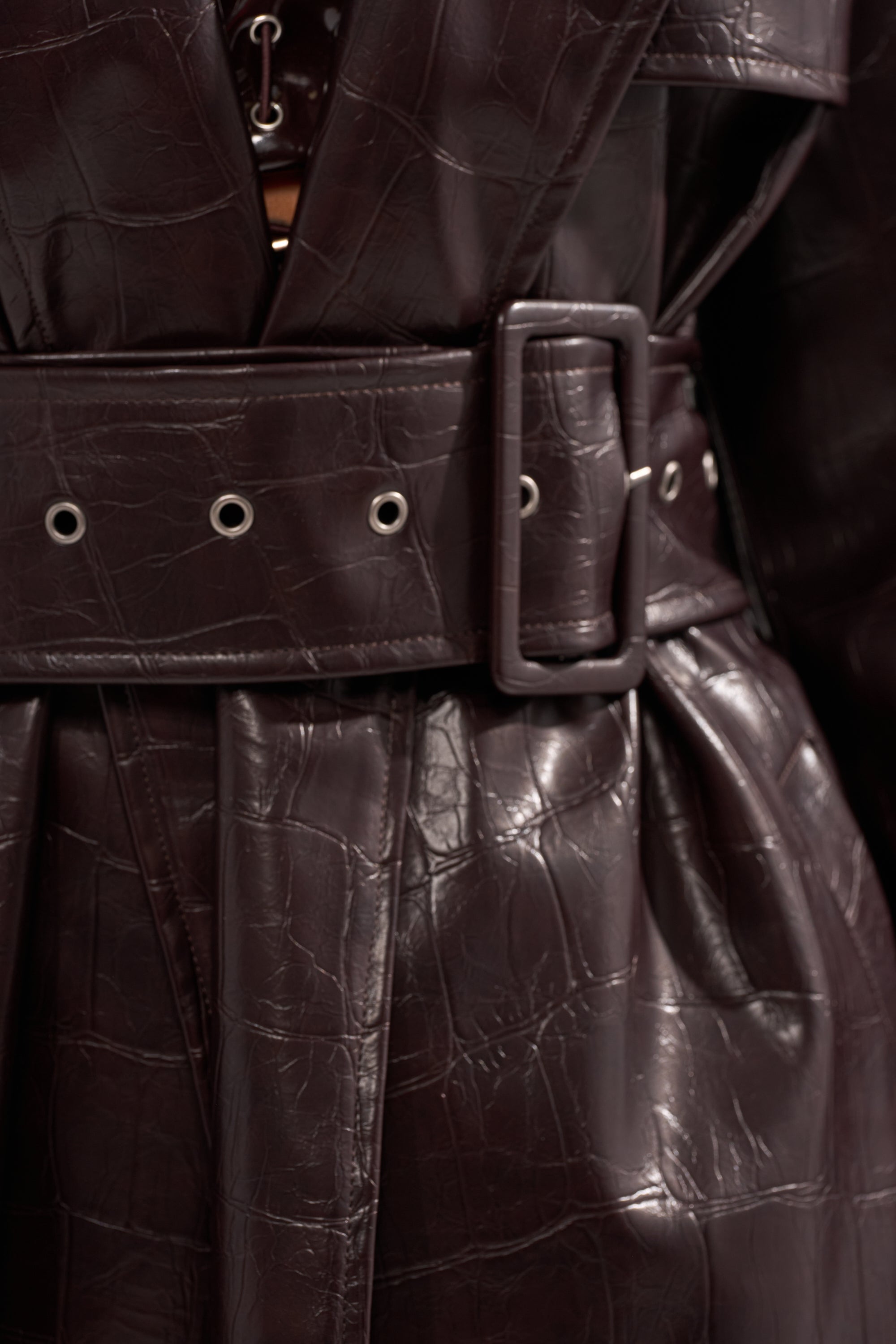 Close-up of the EMBOSSED CROC TRENCH in dark brown, featuring textured faux leather with a crocodile-pattern finish and a wide belt with rectangular buckle and metal eyelets. 9