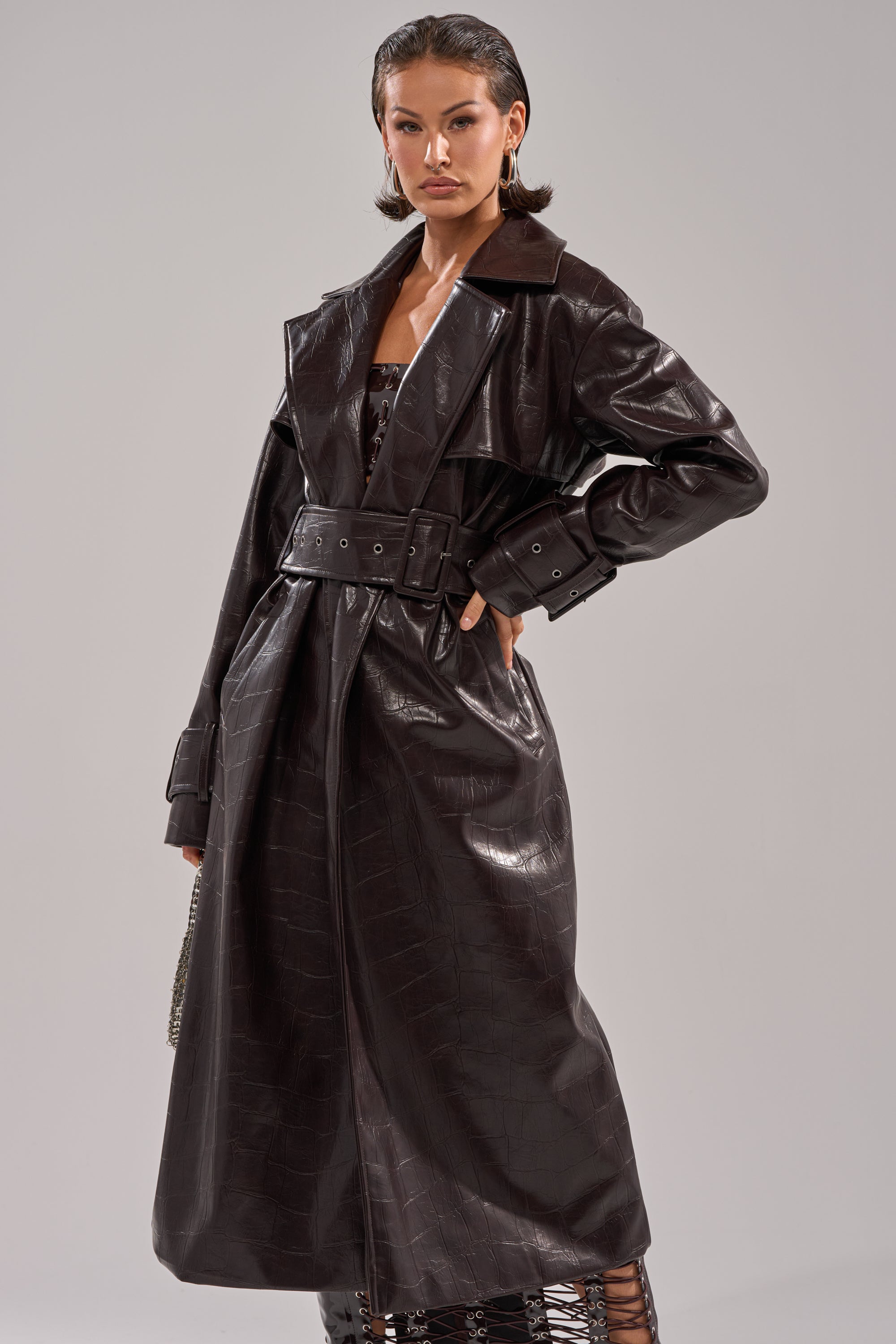A woman exudes confidence in the EMBOSSED CROC TRENCH, a long dark brown crocodile-embossed coat featuring a wide collar, belted waist, and cuffed sleeves. Her short, slicked-back hair and hoop earrings complete the chic look. 1