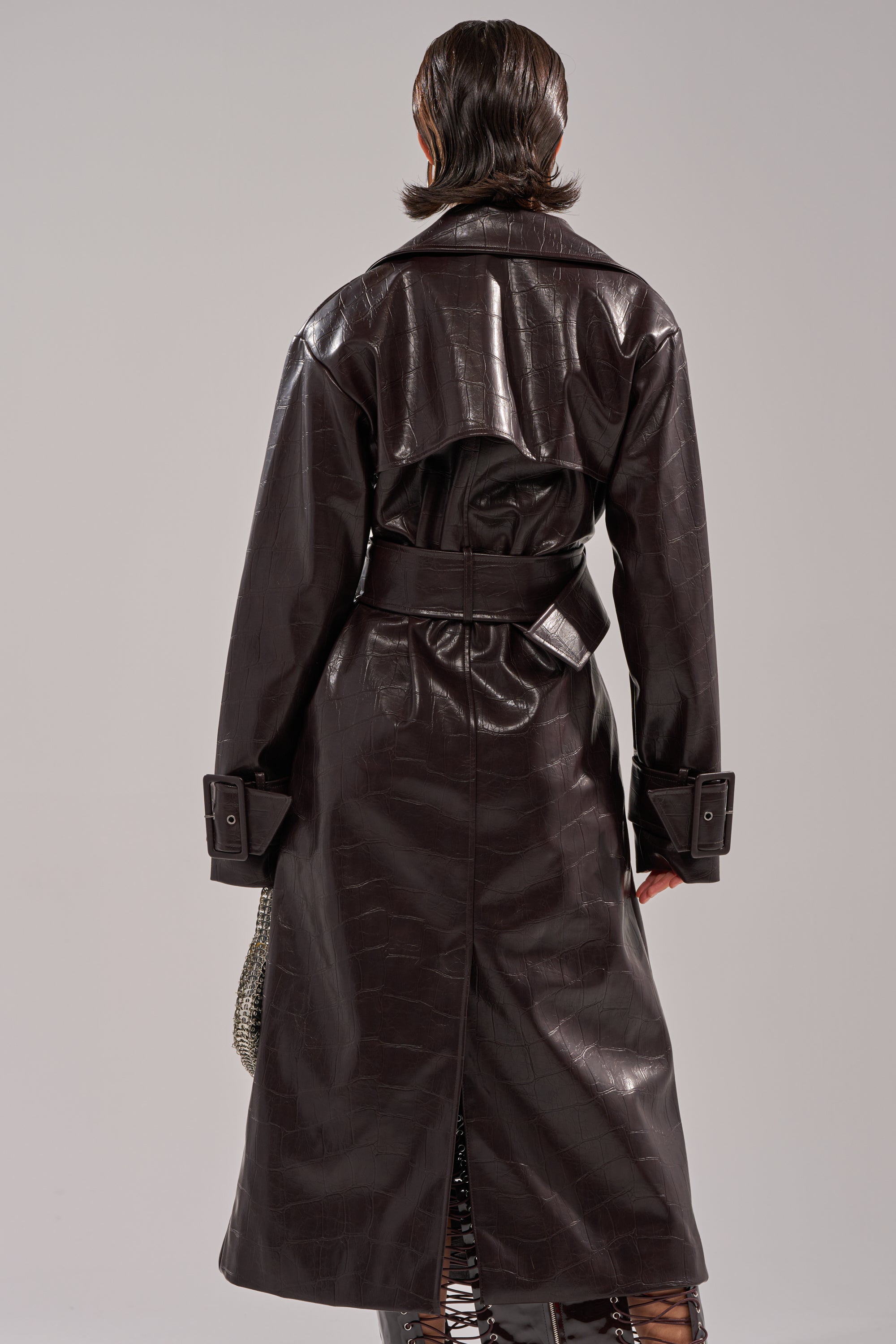 A person faces away, wearing the EMBOSSED CROC TRENCH in dark brown faux leather with a longline fit, belted waist, wide cuffs, embossed croc texture, and back vent against a plain light gray background. 3