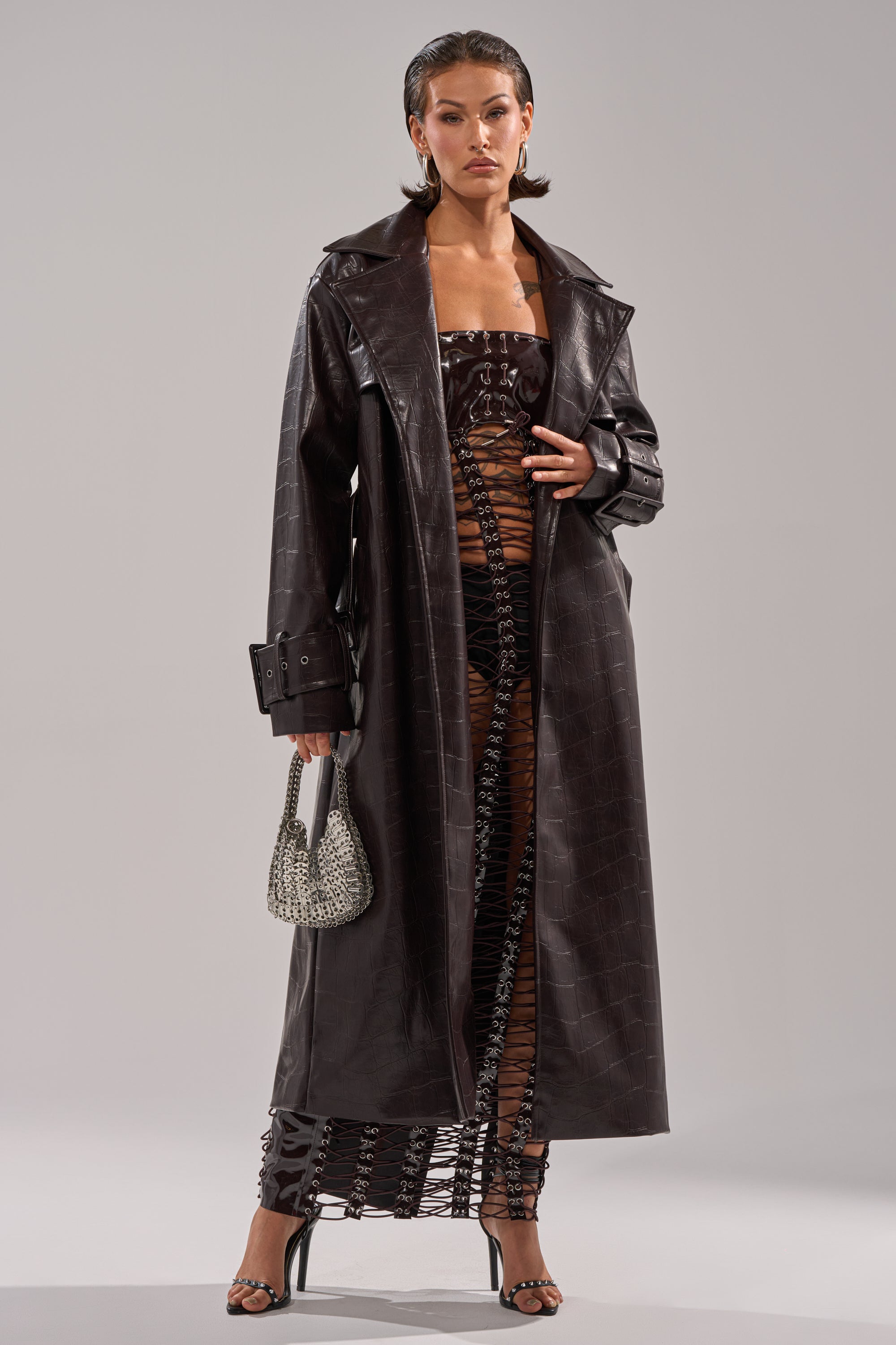 A woman stands confidently in the EMBOSSED CROC TRENCH with a longline silhouette, layered over a black cut-out dress. She carries a small silver handbag, wears strappy heels, and slicks her hair back for a striking finish. 4