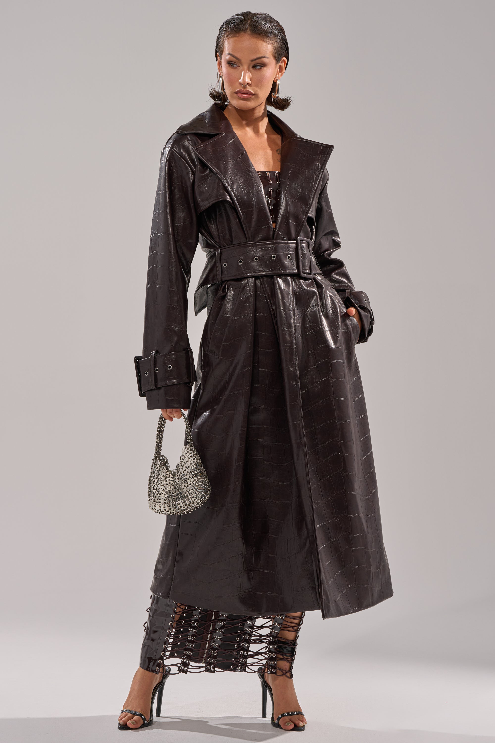 A woman poses in the EMBOSSED CROC TRENCH, a long belted dark brown coat, over a black lace-up dress, paired with a silver beaded handbag and strappy black heels. 5