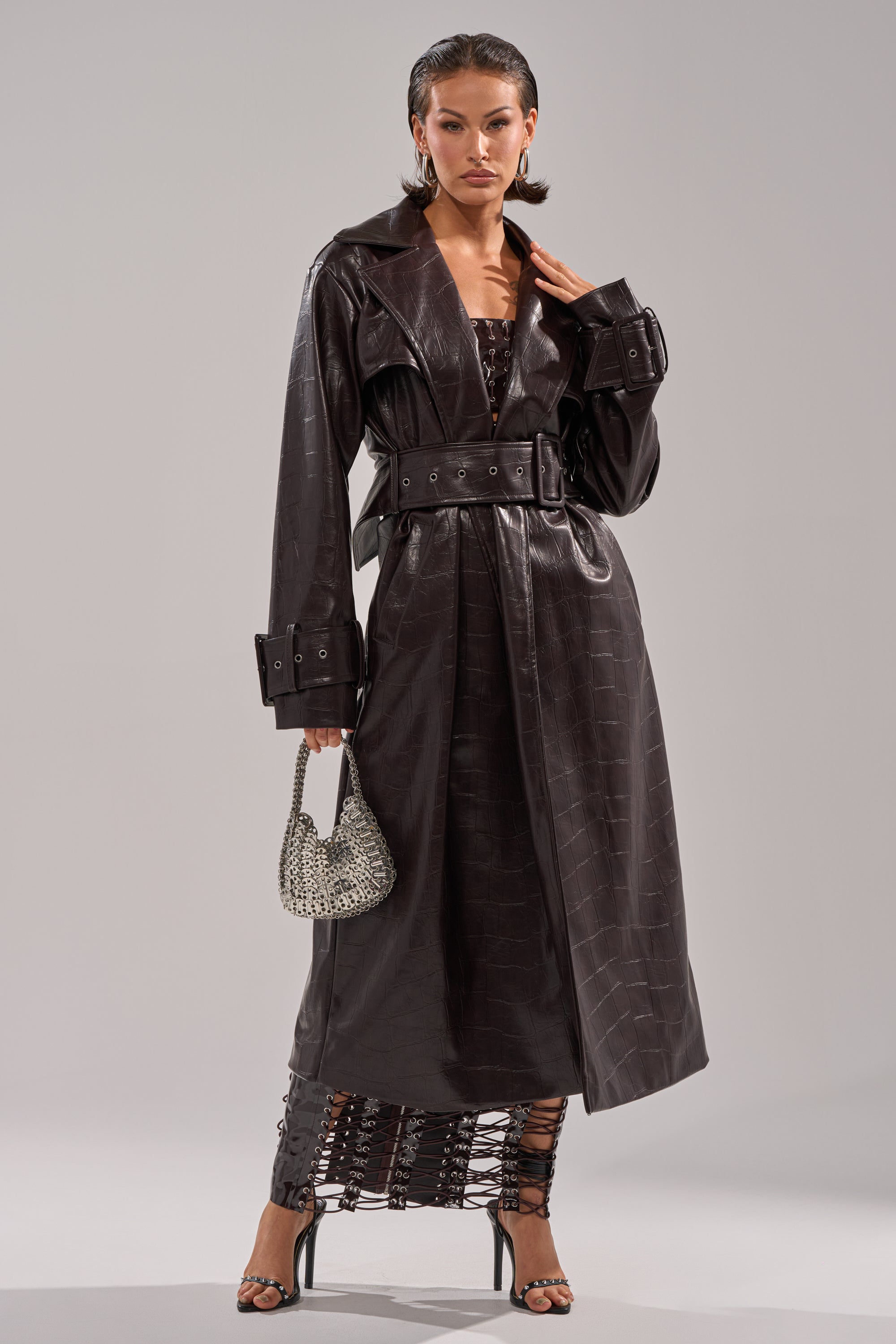 A woman poses in the EMBOSSED CROC TRENCH, a long, belted dark brown crocodile-pattern coat. She holds a silver handbag, wears high-heeled sandals, and reveals a lace-up skirt beneath the coat. 6