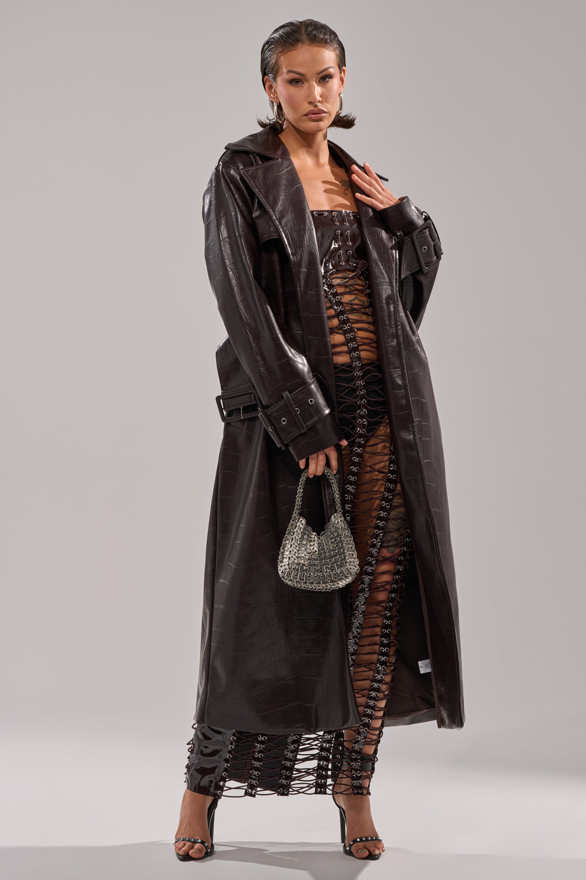 A woman wears the EMBOSSED CROC TRENCH over a matching lace-up dress, carrying a sparkly handbag and wearing open-toed heels. Her slicked-back hair is styled with bold hoop earrings. 7