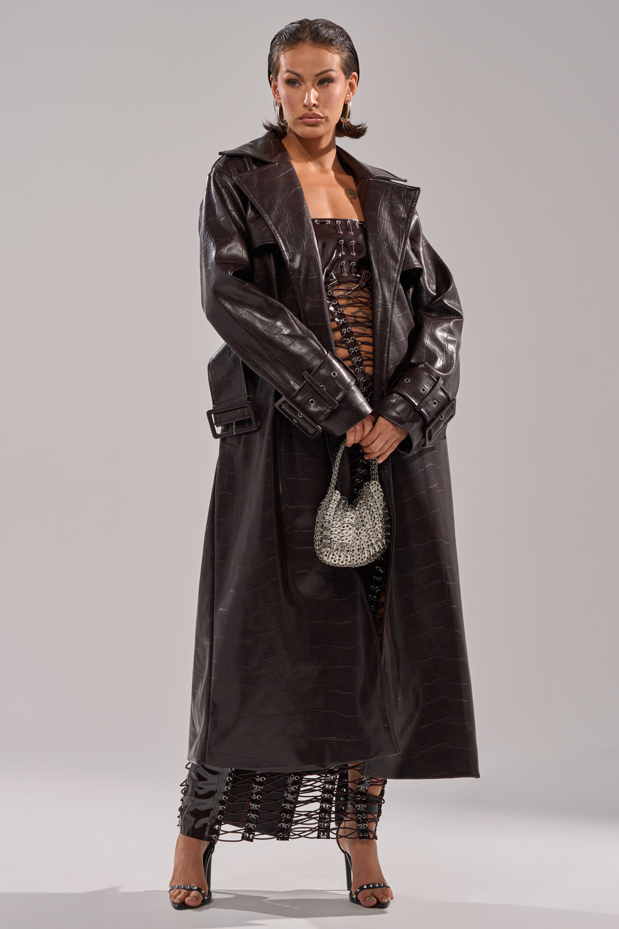 A woman wears the EMBOSSED CROC TRENCH in dark brown over a matching lace-up dress, accessorized with a sparkly silver handbag and black high-heeled sandals. 8