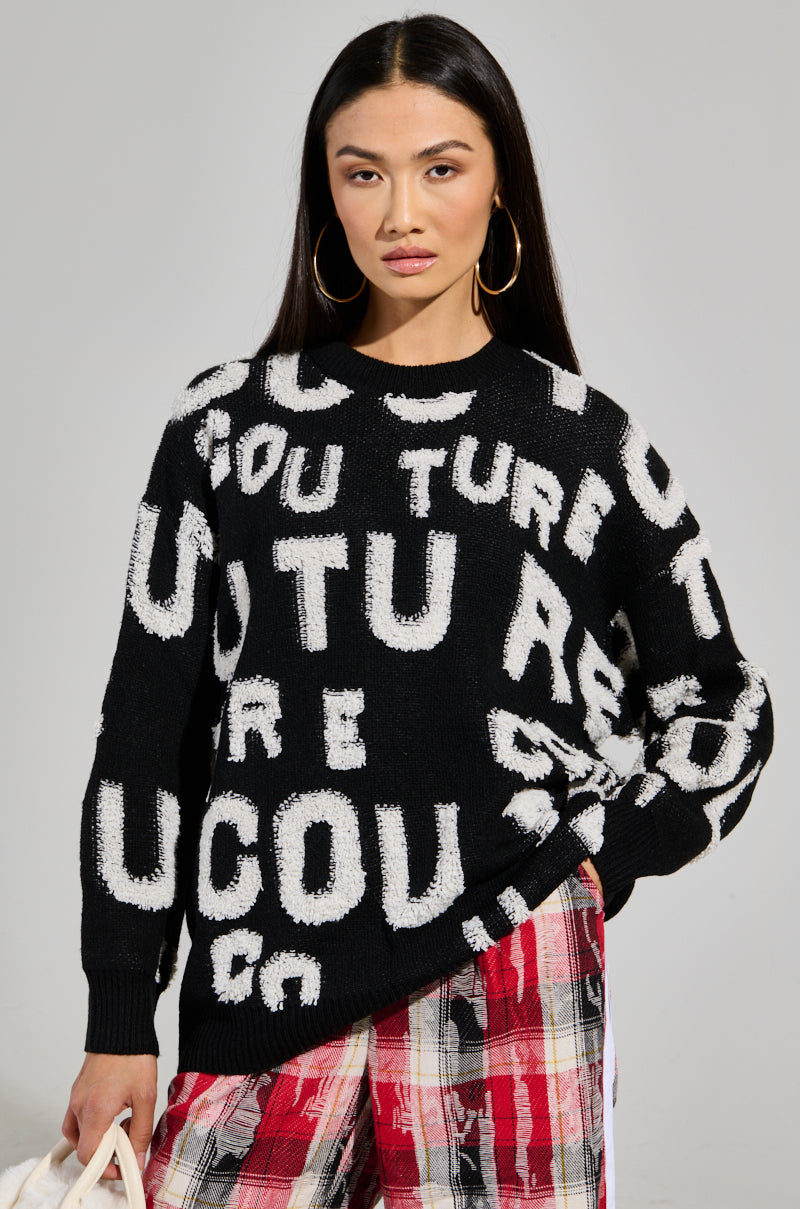 A person with long dark hair is wearing the FOREVER COUTURE LONG SLEEVE KNIT SWEATER, featuring white, abstract letters, and pairs it with red plaid pants. They stand confidently against a neutral background, accessorizing with large hoop earrings and striking a pose with one hand on their hip. 0