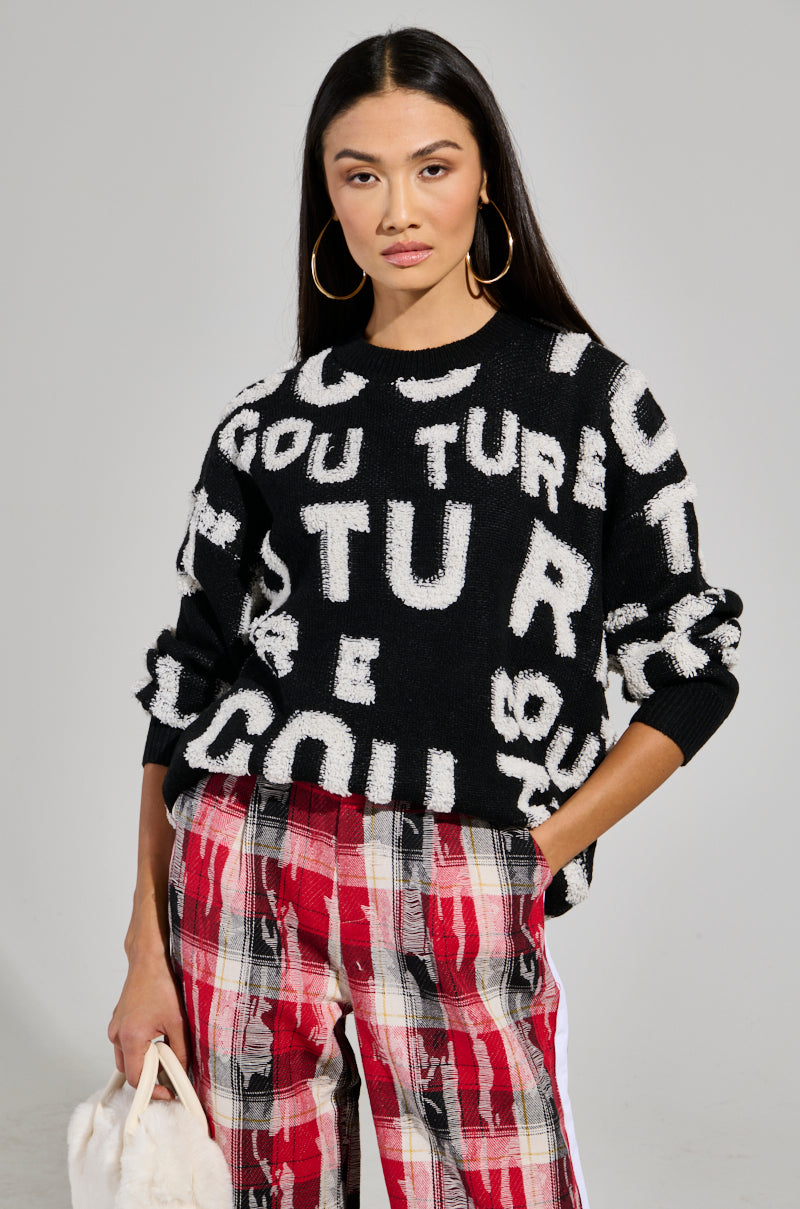 A person with long hair stands confidently, wearing the FOREVER COUTURE LONG SLEEVE KNIT SWEATER in black with bold white lettering, paired with red plaid pants. They hold a white bag and have large hoop earrings, set against a simple gray background. 1