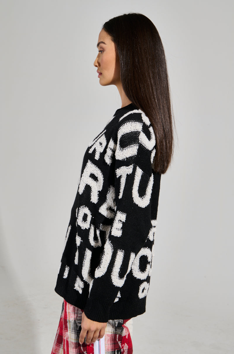 A person with long dark hair is shown in profile against a plain background, wearing the FOREVER COUTURE LONG SLEEVE KNIT SWEATER, which features large, bold white letters, paired with red plaid pants. 2