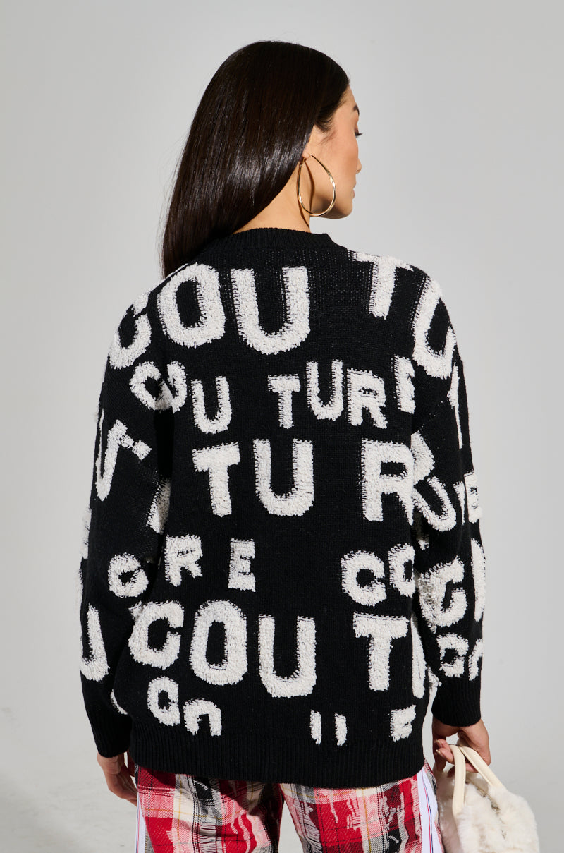 A woman with long dark hair wears the FOREVER COUTURE LONG SLEEVE KNIT SWEATER, showcasing large white letters, paired with red plaid pants. She holds a fluffy white bag. The striking knit design of the sweater enhances the vivid graphic pattern against a neutral background. 3