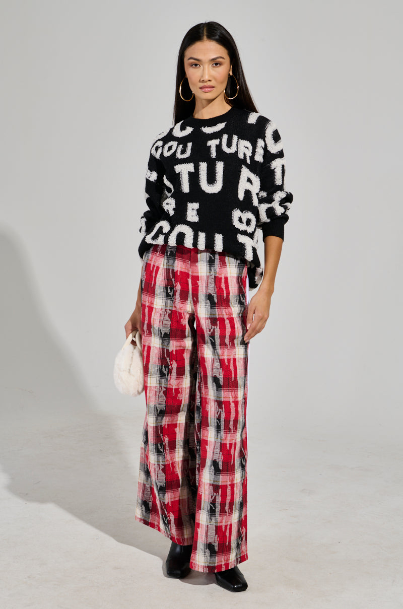 A woman poses against a plain background wearing the FOREVER COUTURE LONG SLEEVE KNIT SWEATER, featuring black and white knit with letters. She pairs it with red and black checkered wide-legged pants and black shoes. Holding a small white fluffy bag, she has long dark hair and hoop earrings. 4