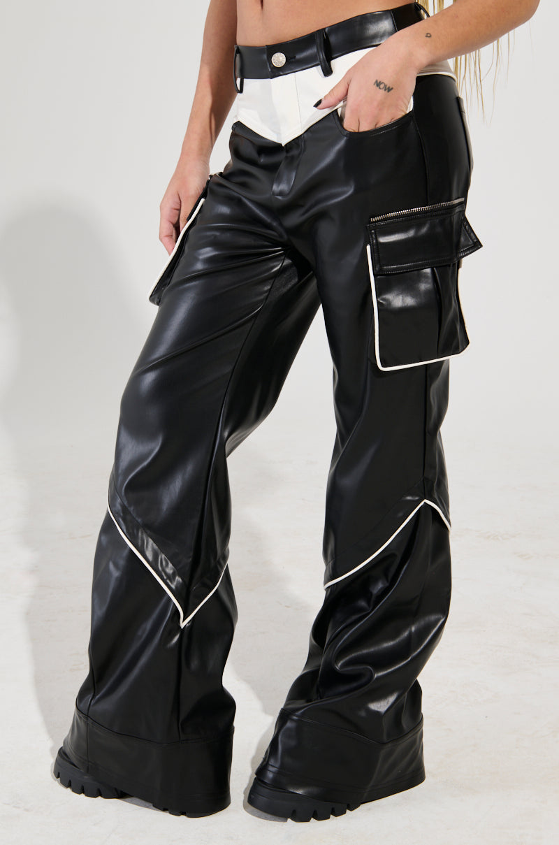 A person stands against a plain background, wearing the THINK AGAIN TROUSER in black and white faux leather featuring cargo flap pockets and white piping. The close-up focuses on the lower half, highlighting the intricate details of these trousers.