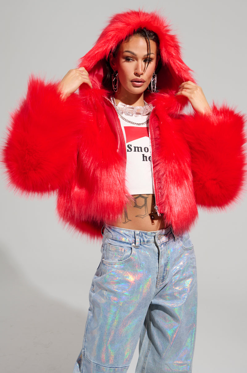 Dressed in the MAEVE HOODED RED FAUX FUR BOMBER, along with holographic jeans and a white crop top, the individual confidently poses against a plain background, gripping the hood with both hands.