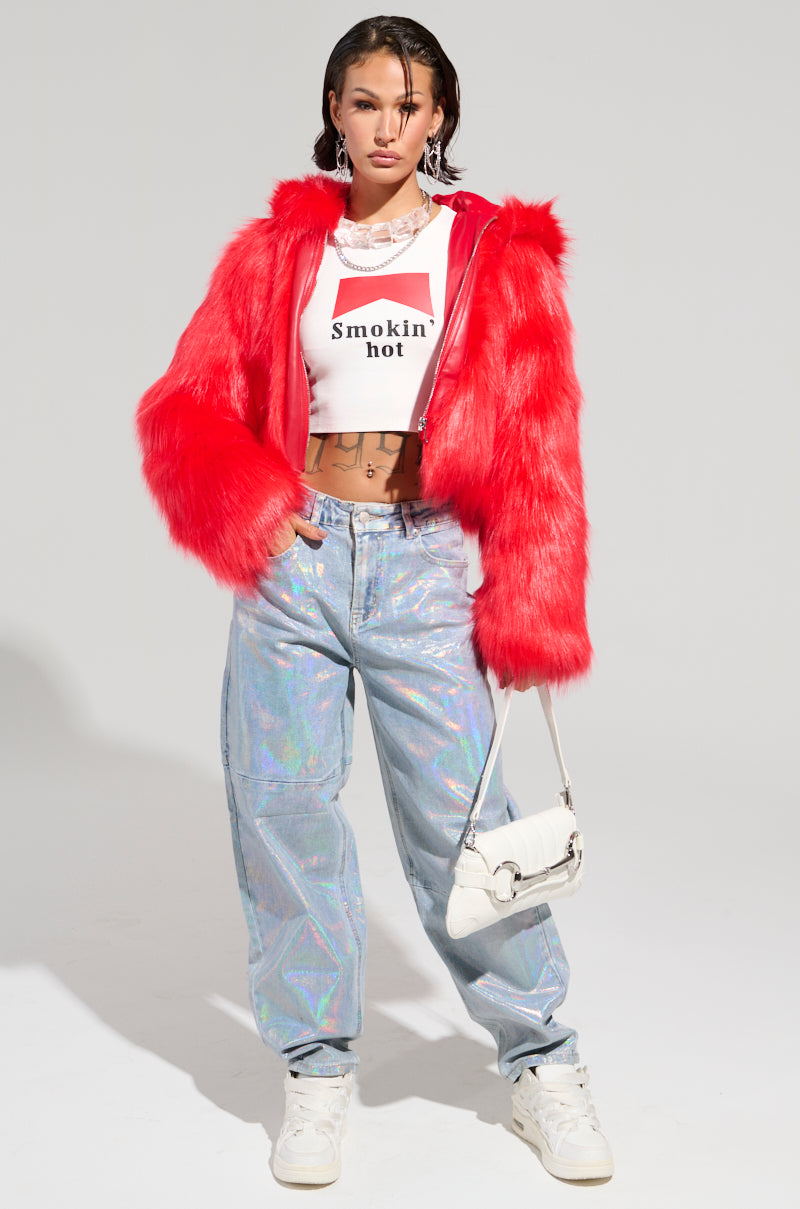 A person stands confidently in the MAEVE HOODED RED FAUX FUR BOMBER, paired with a white crop top emblazoned with "smokin hot," and iridescent pants. They accessorize with a white handbag and wear white sneakers, all set against a plain white background.