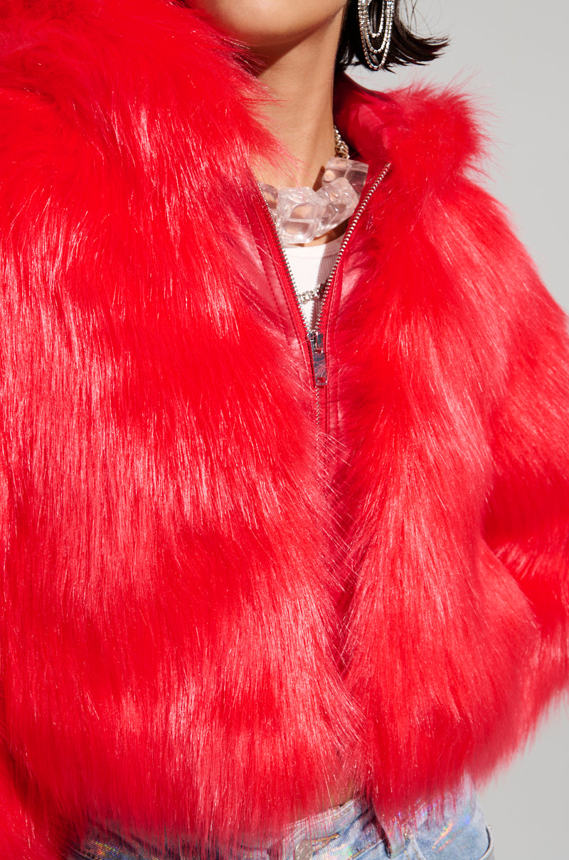 A person wears the MAEVE HOODED RED FAUX FUR BOMBER, complemented by large hoop earrings and a chunky crystal necklace. The jacket's vivid texture is prominently showcased, enhancing the bold and stylish overall look.