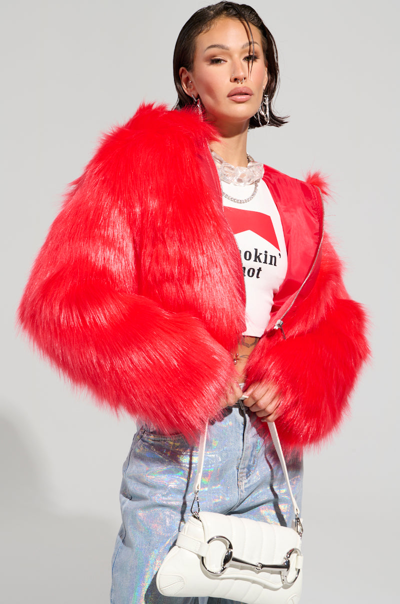 A person wearing the MAEVE HOODED RED FAUX FUR BOMBER layered over a white t-shirt with text, paired with shiny silver jeans. They hold a white handbag and have wet-styled hair. The background is plain gray.
