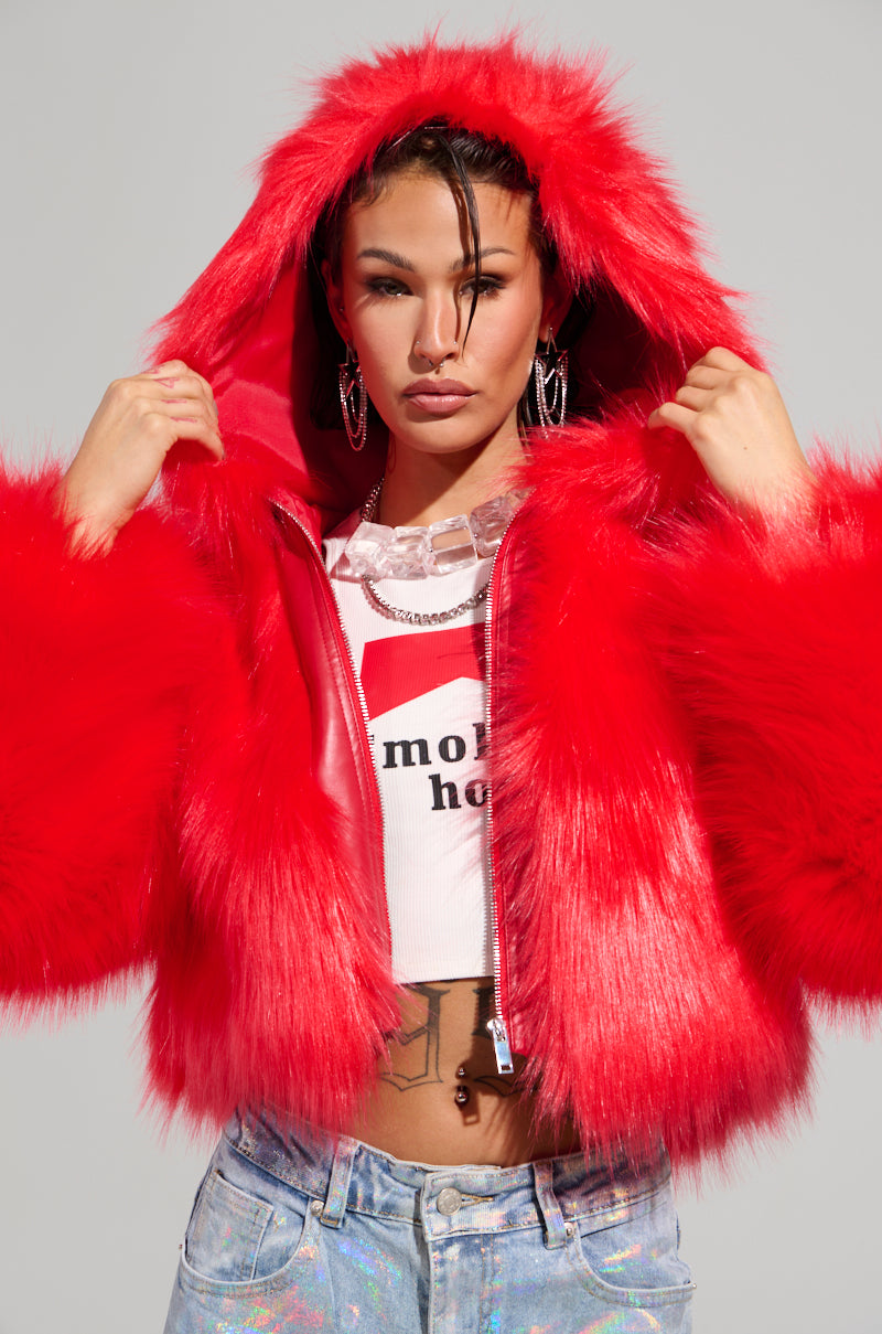 A person is wearing the MAEVE HOODED RED FAUX FUR BOMBER with the hood up, layered over a white shirt with partially visible text. They accessorize with large hoop earrings and stand against a neutral background.