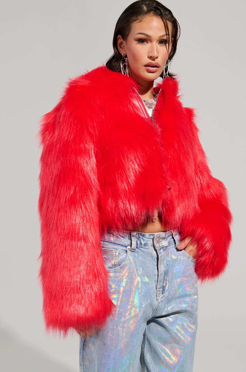A person wearing the MAEVE Hooded Red Faux Fur Bomber and light blue iridescent jeans stands against a plain background. The oversized jacket with long sleeves complements their shoulder-length hair, while large silver earrings add a touch of elegance.
