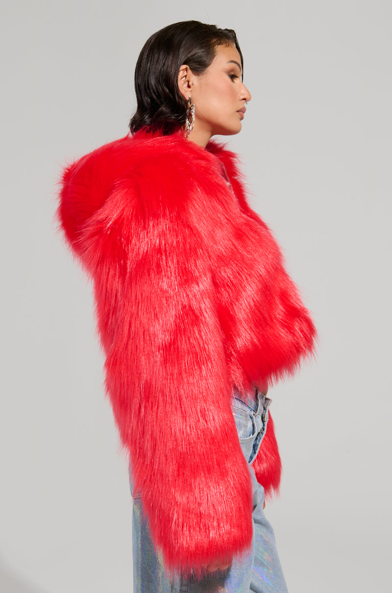 A person wearing the MAEVE Hooded Red Faux Fur Bomber coat and dangling earrings is facing sideways against a plain gray background. The coat's hood adds flair to their ensemble of blue jeans.