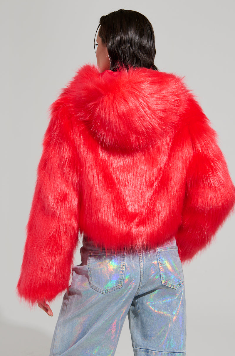 A person wearing the MAEVE HOODED RED FAUX FUR BOMBER, which features a vibrant red hue and a hood, stands facing away. They complement the bright fur with holographic jeans that reflect various colors, creating a shiny contrast against the bomber. The background is a plain grey.