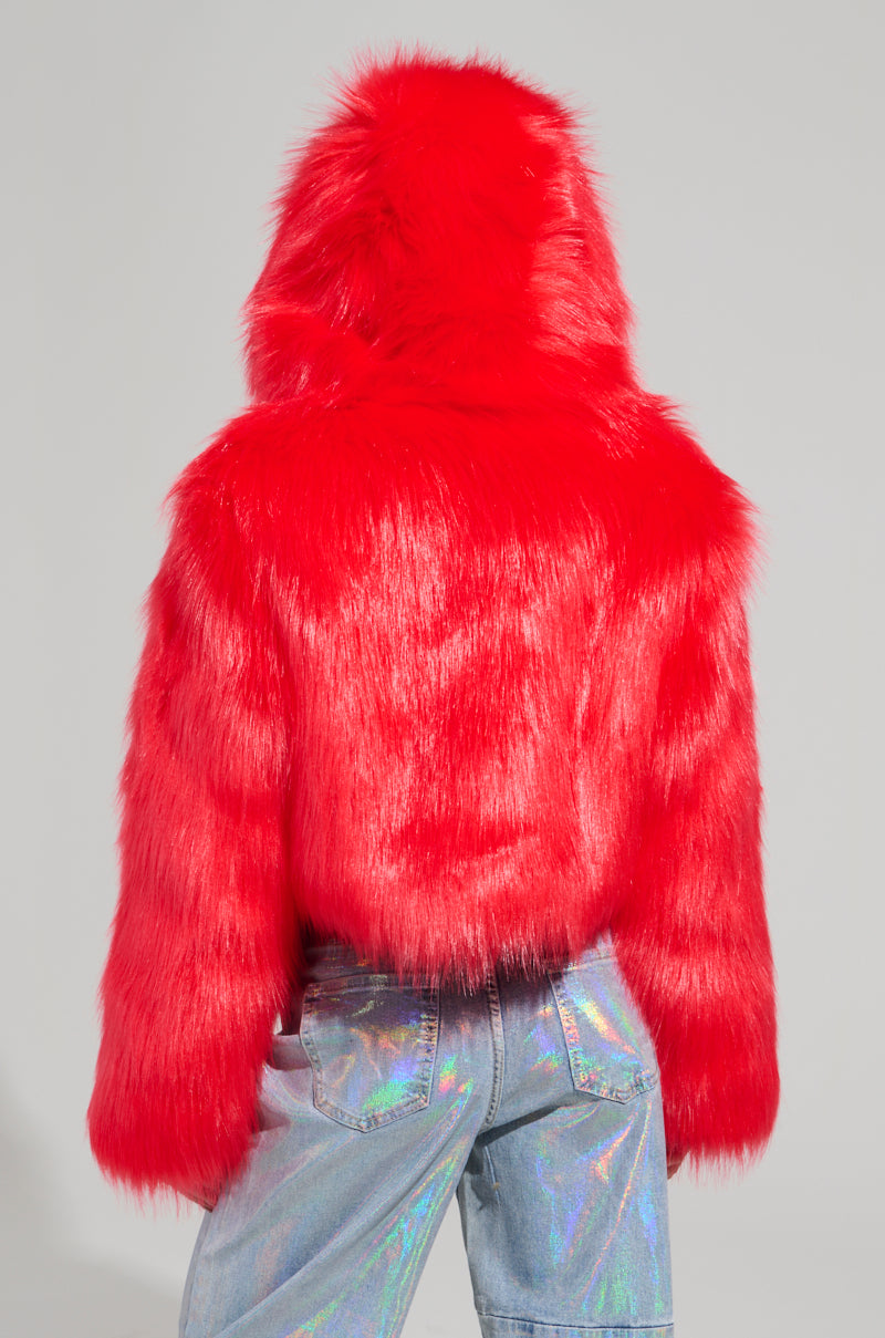 A person wearing the MAEVE Hooded Red Faux Fur Bomber stands facing away. The jacket is glossy and eye-catching, perfectly paired with iridescent jeans that create a rainbow effect in the light. Against a neutral background, they make a captivating style statement.