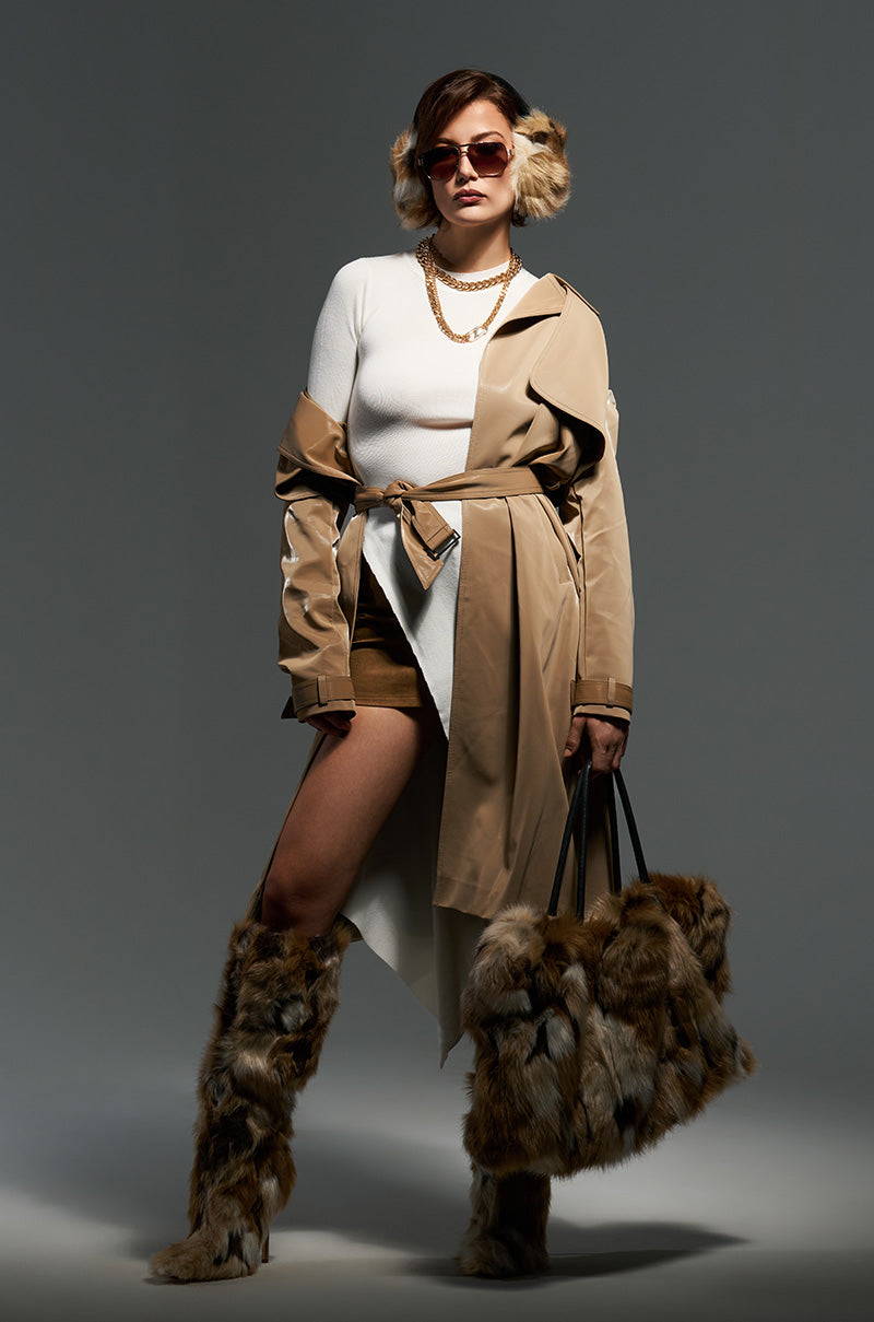 Dressed in the LEAH LO LIQUID VELVET TRENCH, a person exudes confidence with its light brown hue and maxi-length silhouette. They've paired it with a white top, brown shorts, and fur boots for a chic ensemble. Sunglasses, hoop earrings, and a chain necklace add stylish accents as they confidently hold a fur bag against the neutral gray backdrop.