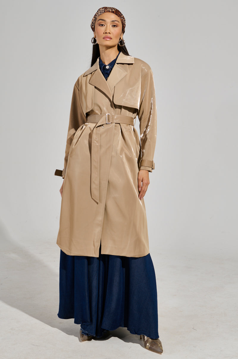 Confidently, a person wears the LEAH LO Liquid Velvet Trench over a long, dark blue dress. Their look is complemented by a patterned headband and shiny shoes, set against a plain light gray background.