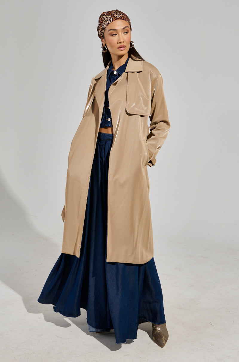 A stylish person poses confidently in the LEAH LO LIQUID VELVET TRENCH over a dark blue outfit with a leopard-print headscarf. They stand against a plain background, exuding modern elegance with their maxi-length silhouette.