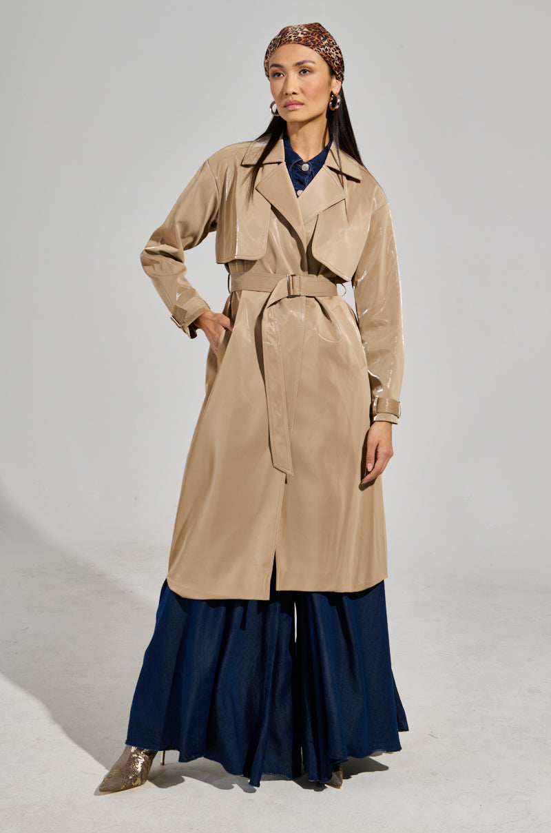 A person wearing the LEAH LO LIQUID VELVET TRENCH with a belt stands confidently in a neutral-colored space, sporting a dark blue outfit and a patterned headband. One hand rests on their hip while the other hangs by their side.