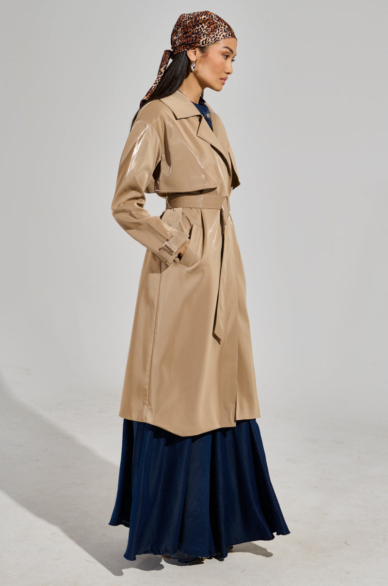 Wearing the chic LEAH LO LIQUID VELVET TRENCH with its elegant beige maxi-length design, a person stands sideways dressed in a long dark blue gown. They have accentuated their look with a patterned headscarf tied around their head, beautifully set against the light gray backdrop.