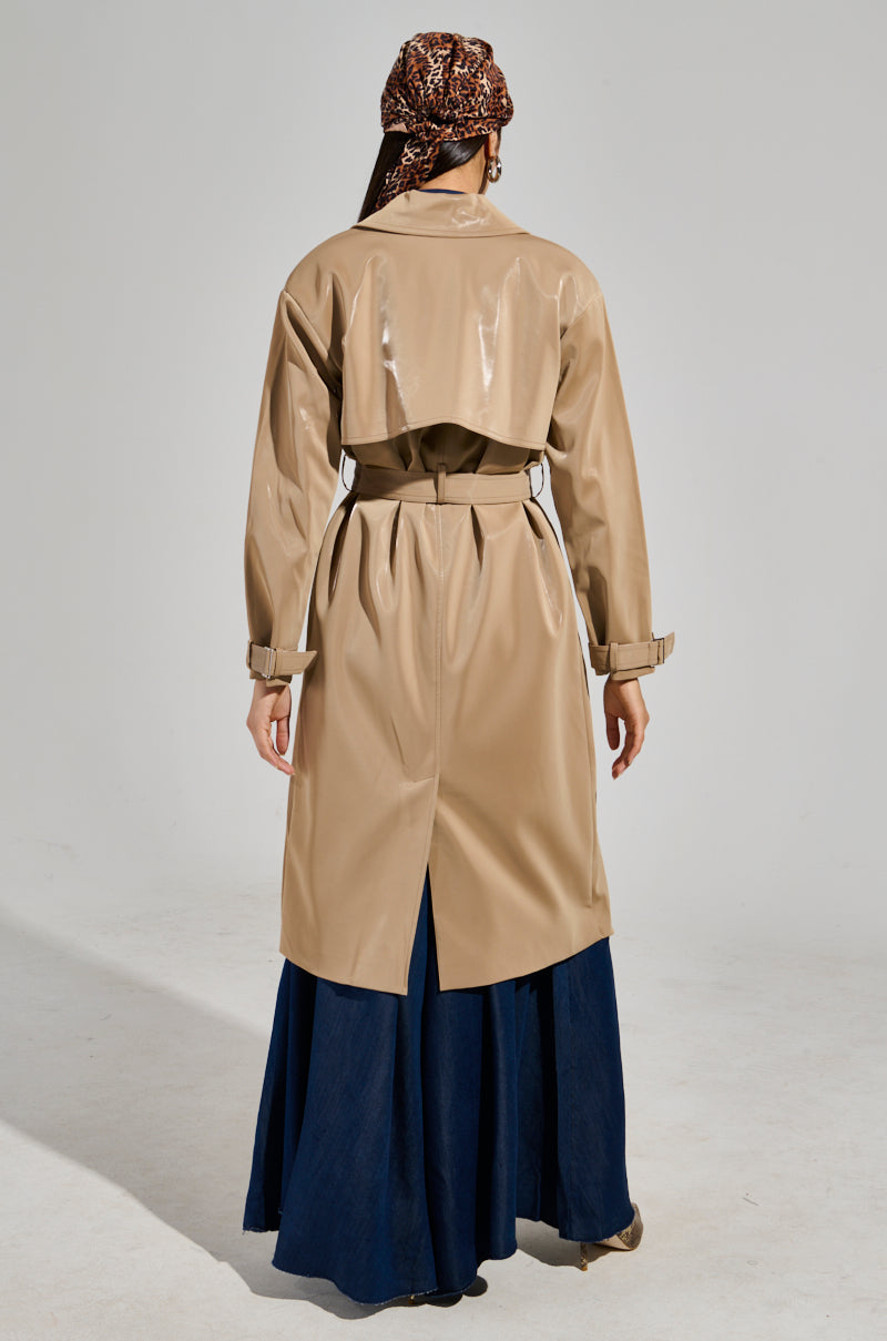 A person is wearing the LEAH LO LIQUID VELVET TRENCH in tan, featuring a maxi-length silhouette, paired with a patterned headscarf. They are standing with their back to the camera, revealing a dark blue skirt peeking from below the coat. The background is a plain off-white color.