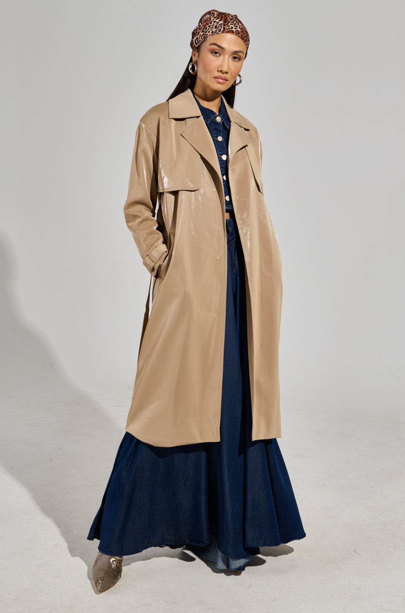 A person stands confidently in the LEAH LO LIQUID VELVET TRENCH, which drapes elegantly over a maxi-length silhouette, with a patterned scarf on their head, all set in a vibrant studio backdrop.