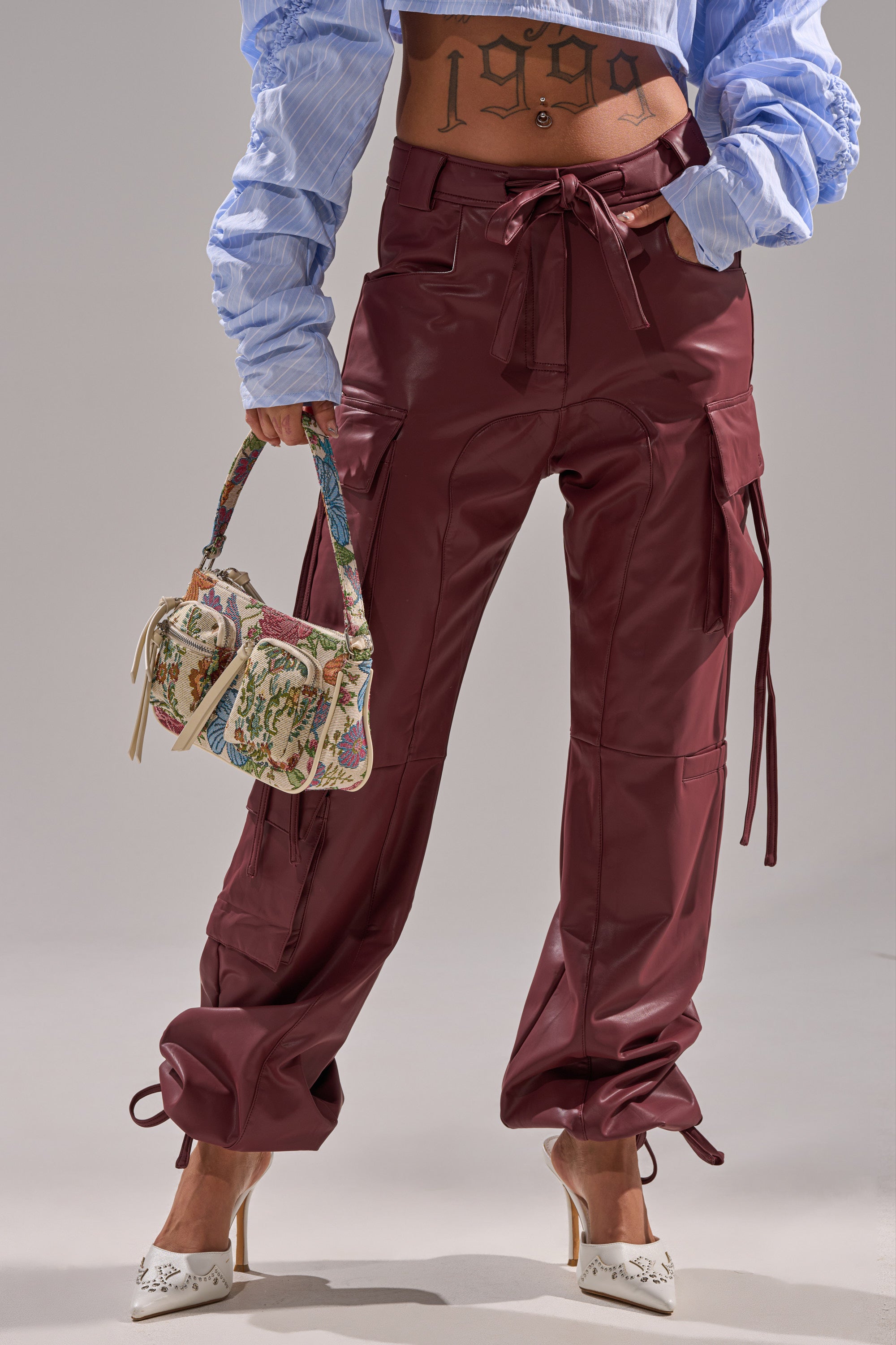 Wearing the DO SOMETHING CRAZY CARGO TROUSER in high-waisted burgundy faux leather with a wide-leg fit, paired with a blue cropped blouse, white heeled shoes, and a floral handbag. A "1999" tattoo is visible on their torso.