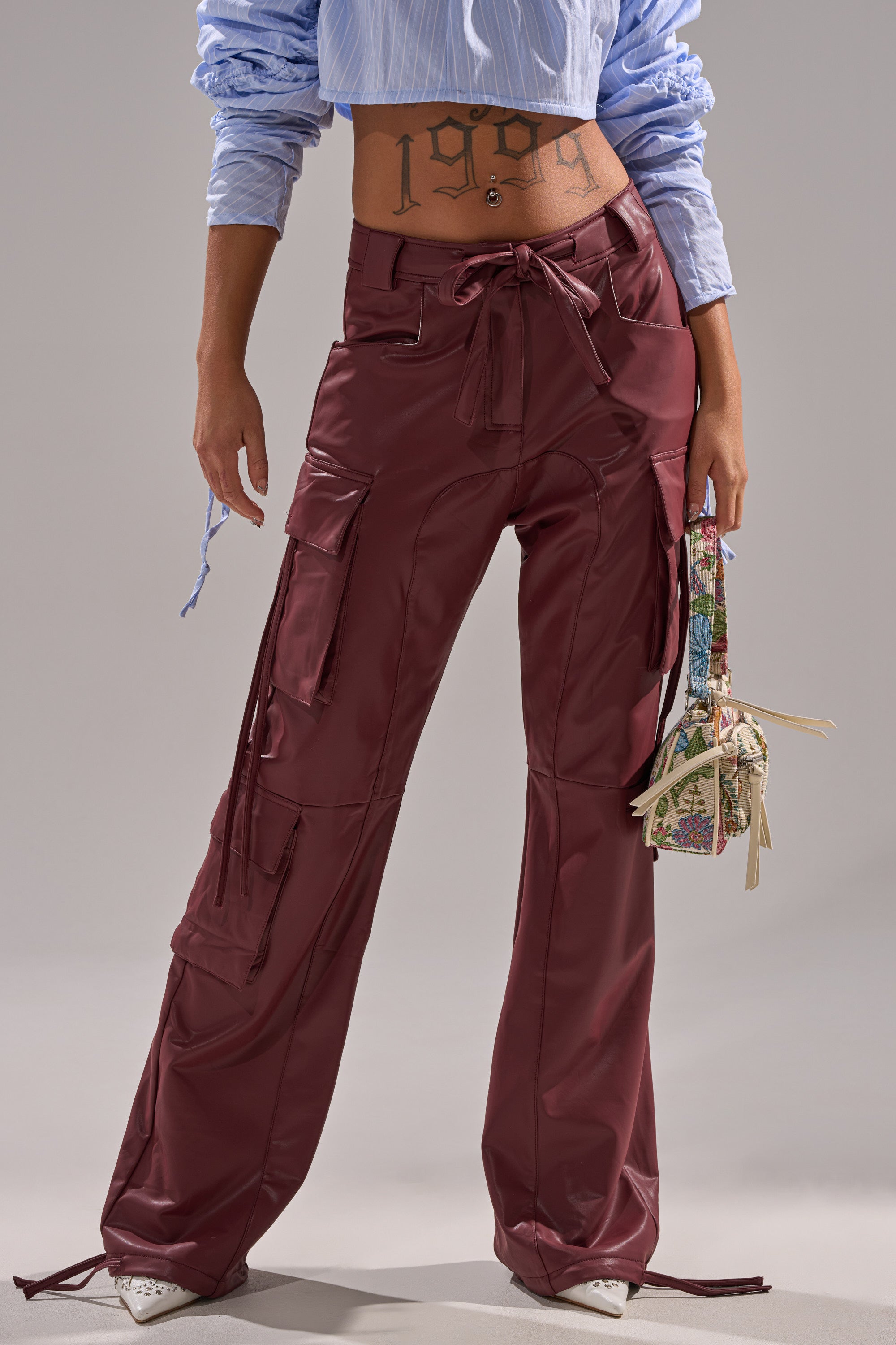 Wearing DO SOMETHING CRAZY CARGO TROUSER in shiny maroon, a blue cropped shirt with ruched sleeves, white heels, and holding a small colorful handbag, with "1999" tattoo visible on the lower abdomen.