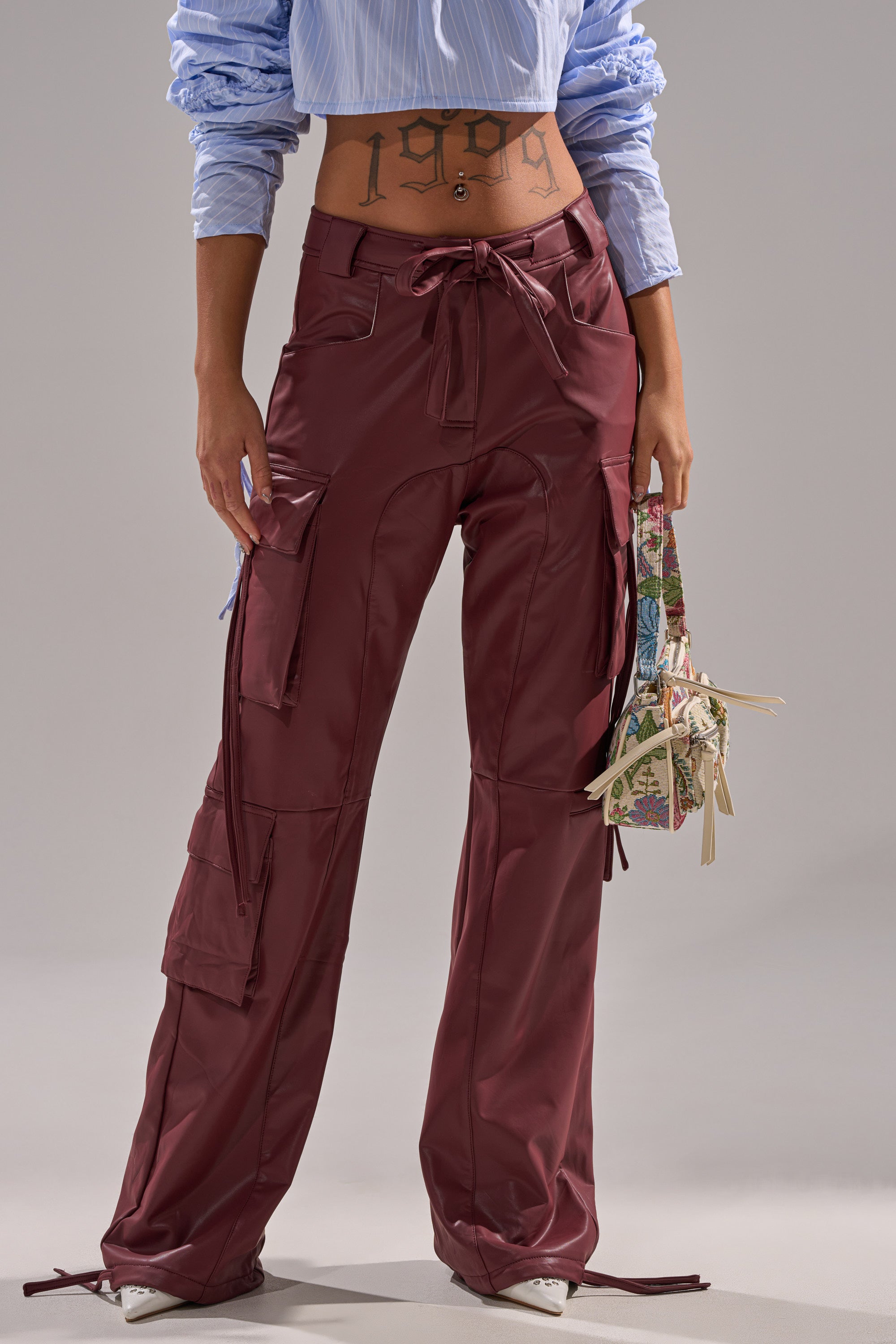 Wearing DO SOMETHING CRAZY CARGO TROUSER in burgundy with a wide-leg fit, a blue cropped shirt with rolled-up sleeves, and holding a small floral handbag, their midriff is exposed to show a "1999" tattoo.