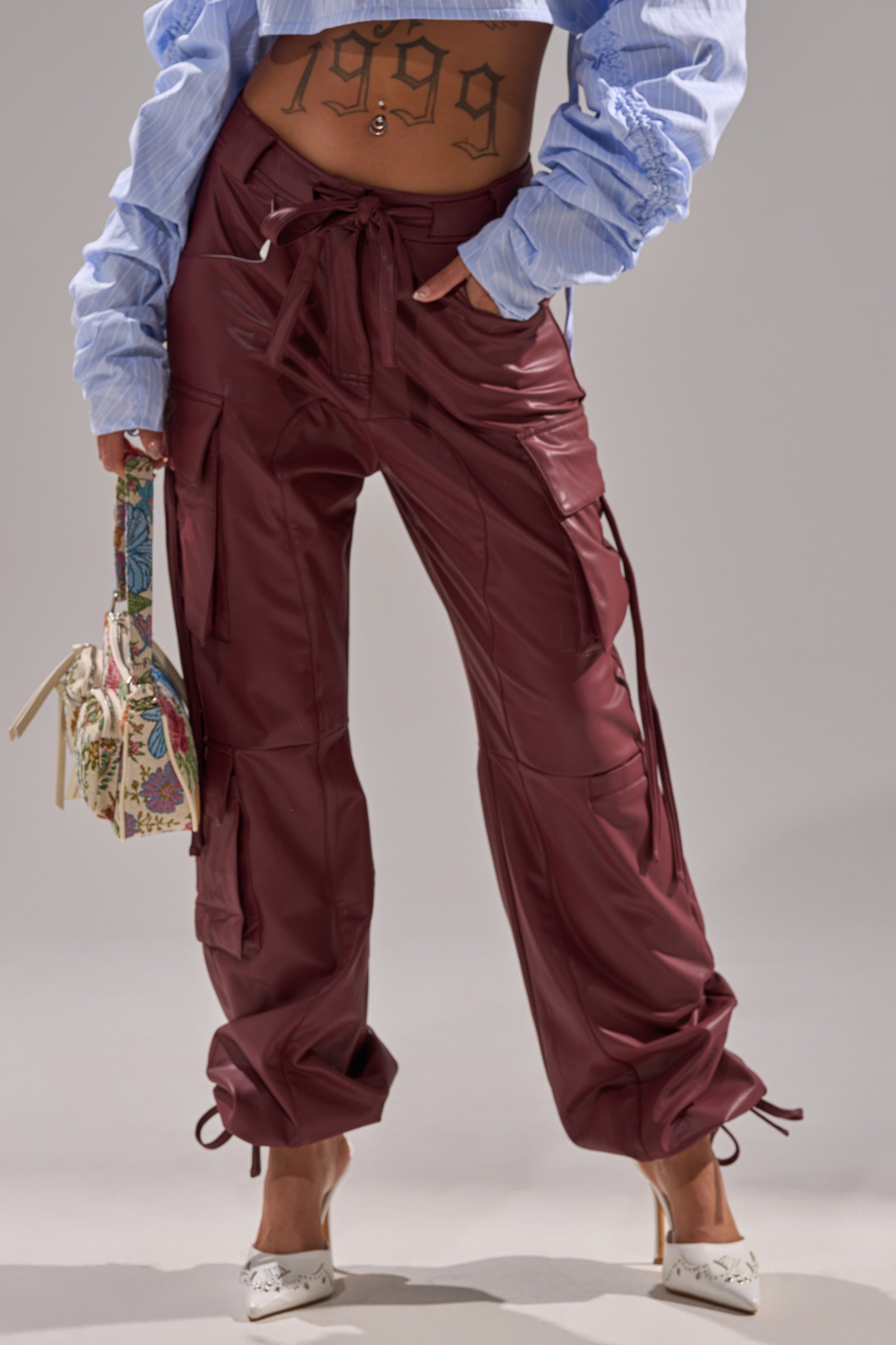 Someone wearing the DO SOMETHING CRAZY CARGO TROUSER in burgundy, a cropped blue shirt revealing a "1999" tattoo, white heels, and holding a patterned handbag stands against a plain background.