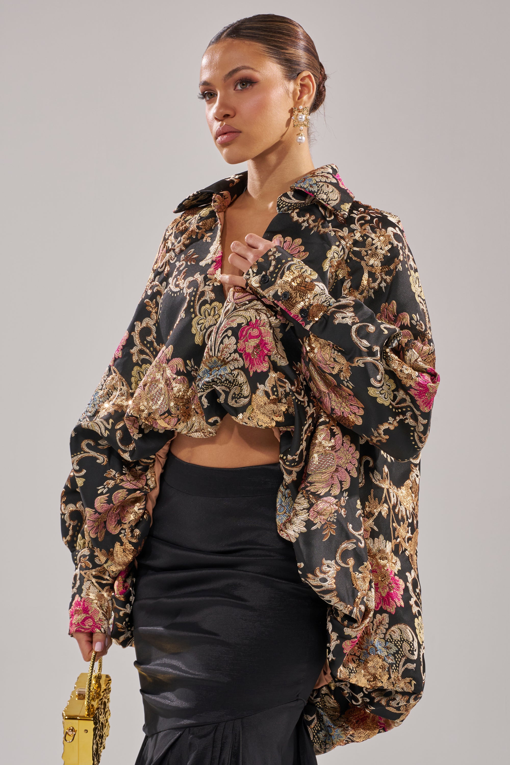 A woman poses in the HIGH SOCIETY HIGH LOW BROCADE BLOUSE—a gold-tone metallic top with floral patterns—paired with a black satin skirt, gold earrings, and a small gold bag, her hair tied back.
