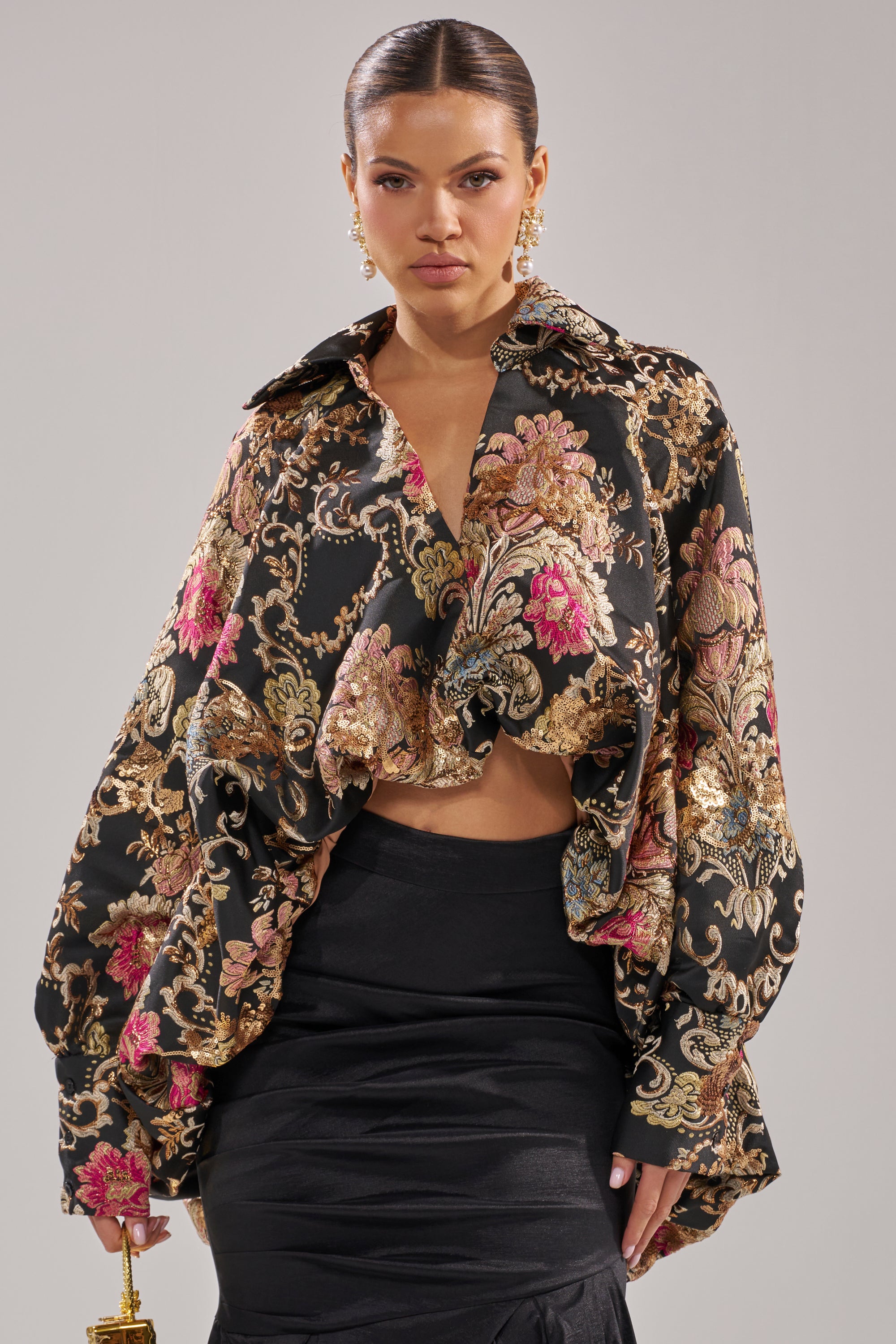 A woman stands against a plain light gray background wearing the HIGH SOCIETY HIGH LOW BROCADE BLOUSE with gold and pink floral patterns, paired with a high-waisted black skirt. She accessorizes with a small gold purse and gold earrings.