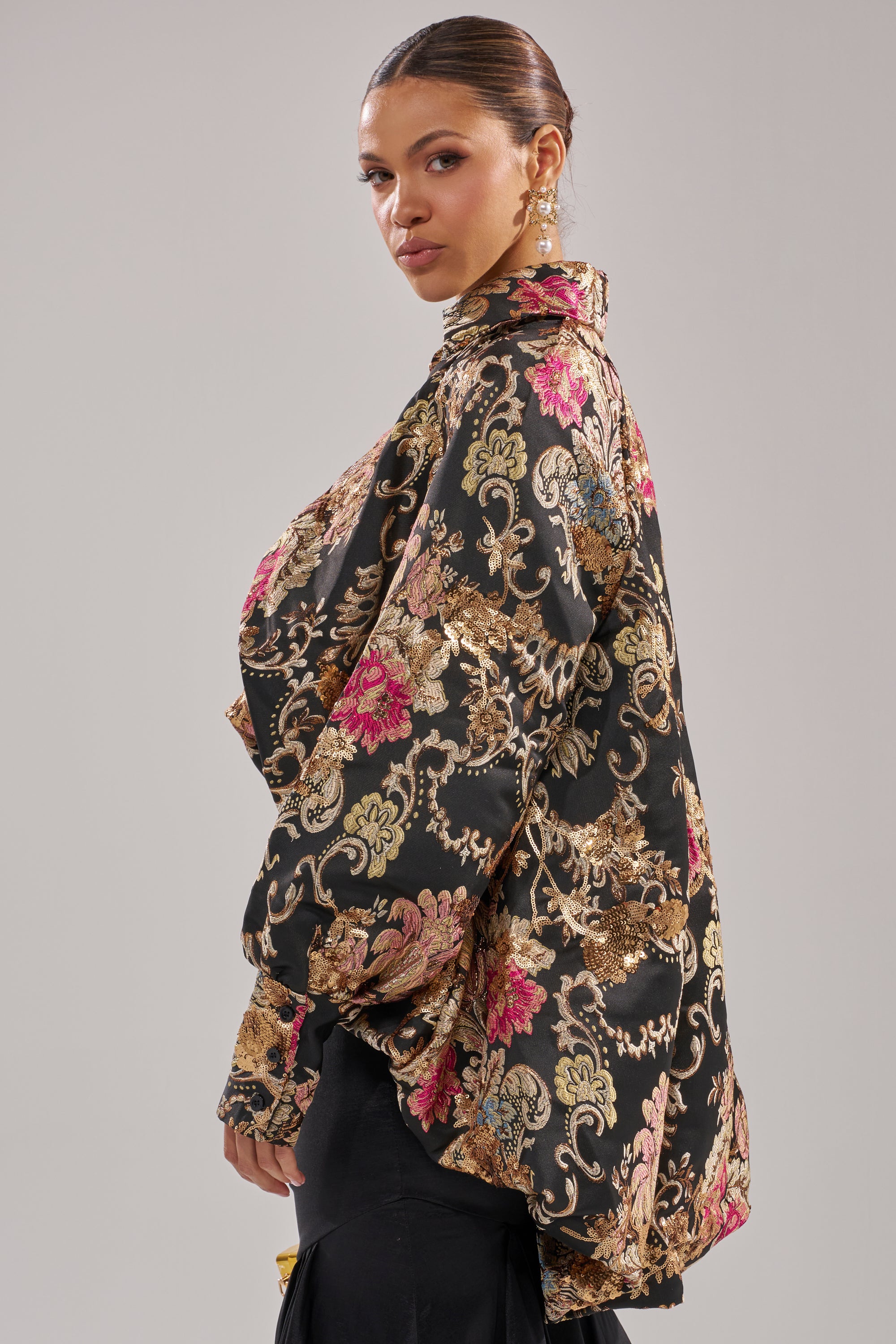 A woman poses in the HIGH SOCIETY HIGH LOW BROCADE BLOUSE, featuring a loose black and gold floral design with pink accents over sequins. With her hair in a low bun and dangling earrings, she stands against a light gray background.