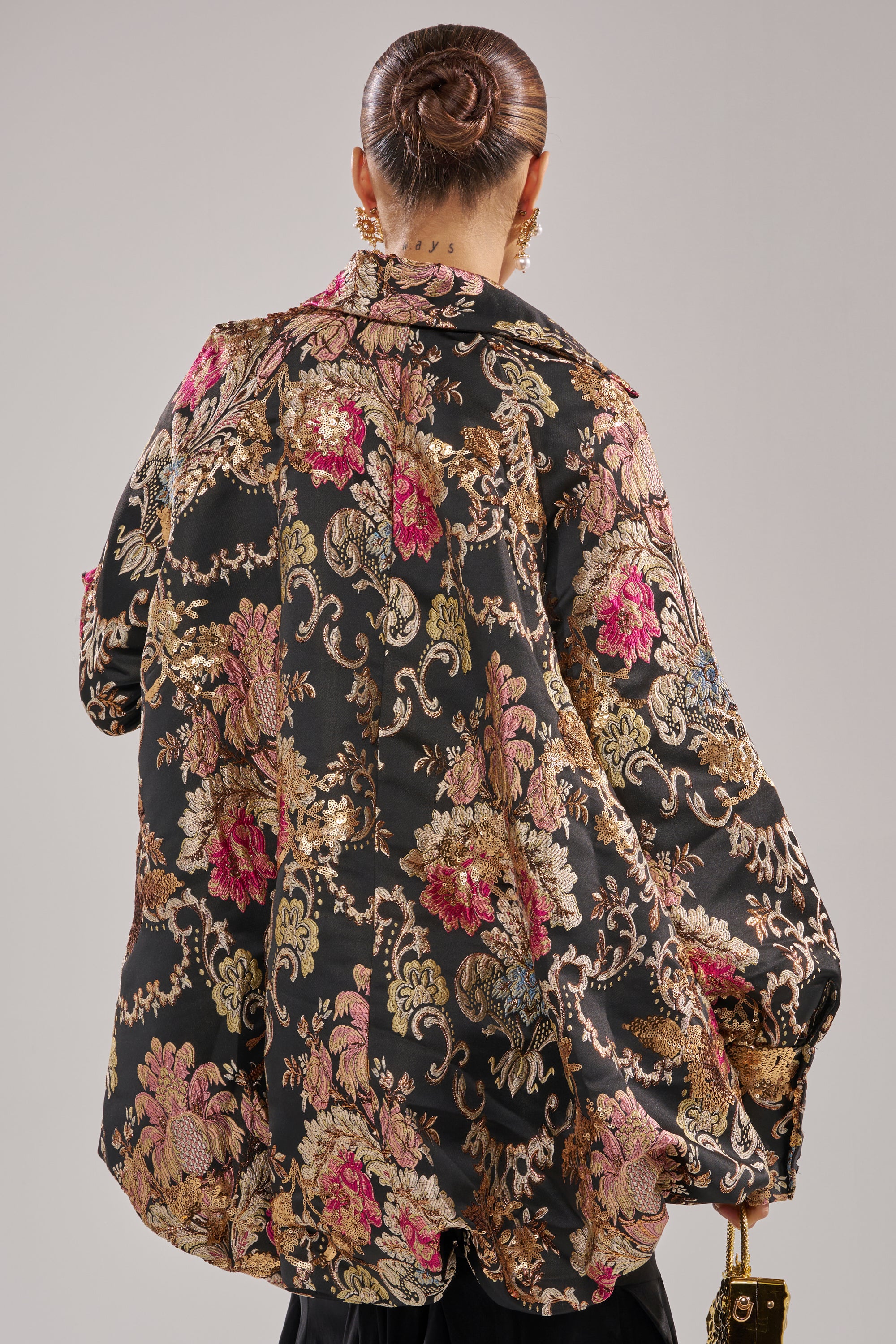 A person with a neat bun is seen from the back, wearing the HIGH SOCIETY HIGH LOW BROCADE BLOUSE—a black, gold, and pink oversized floral-patterned top with metallic detailing—and holding a small handbag.
